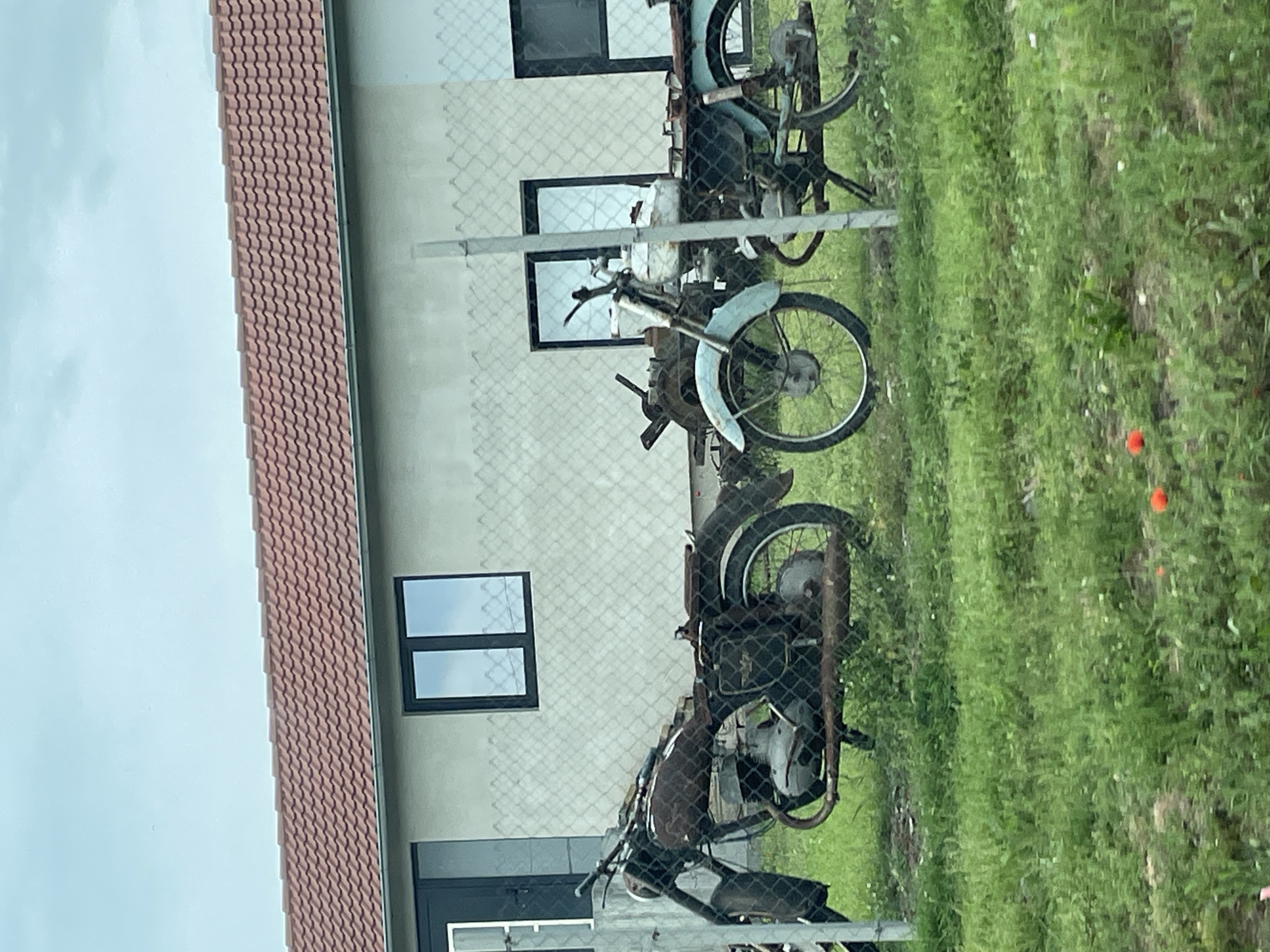 The image depicts a scene in an outdoor setting. Three motorcycles, appearing to be in a state of disrepair, are visible behind a chain-link fence. Behind the fence, a building with a tiled roof and several windows is present. The surrounding environment consists of green grass. The image was presumably taken in Ushintsi, Bulgaria. There are no people present in the image.