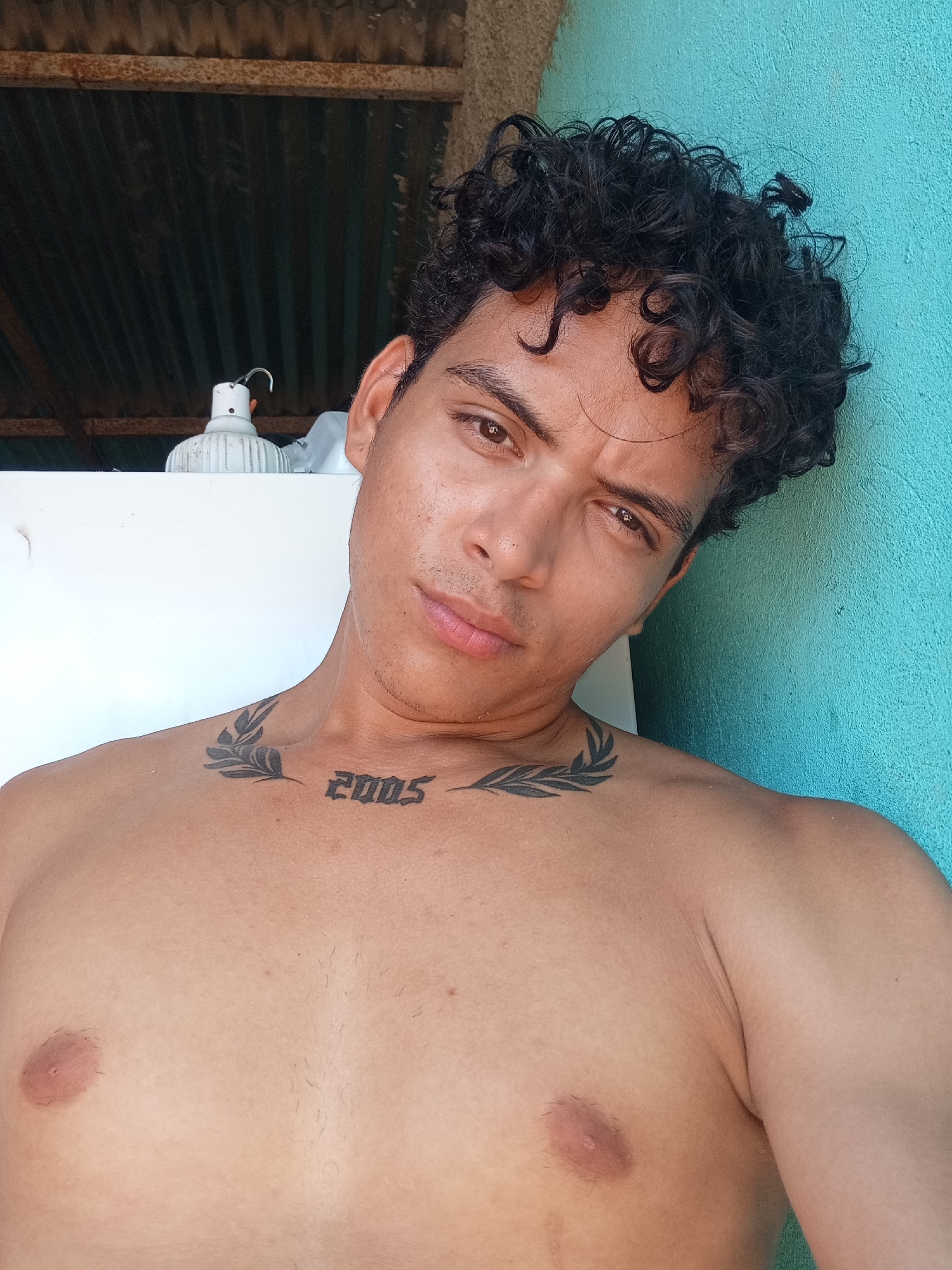 A young adult male with curly dark hair and a tattoo on his chest is leaning against a turquoise wall. He is shirtless and has a relaxed expression. In the background, a corrugated metal roof is visible, supported by a rusty metal beam. A white object, possibly a light fixture, is also partially visible. The scene appears to be outdoors, likely in a rural or semi-rural setting in Altagracia, Venezuela.