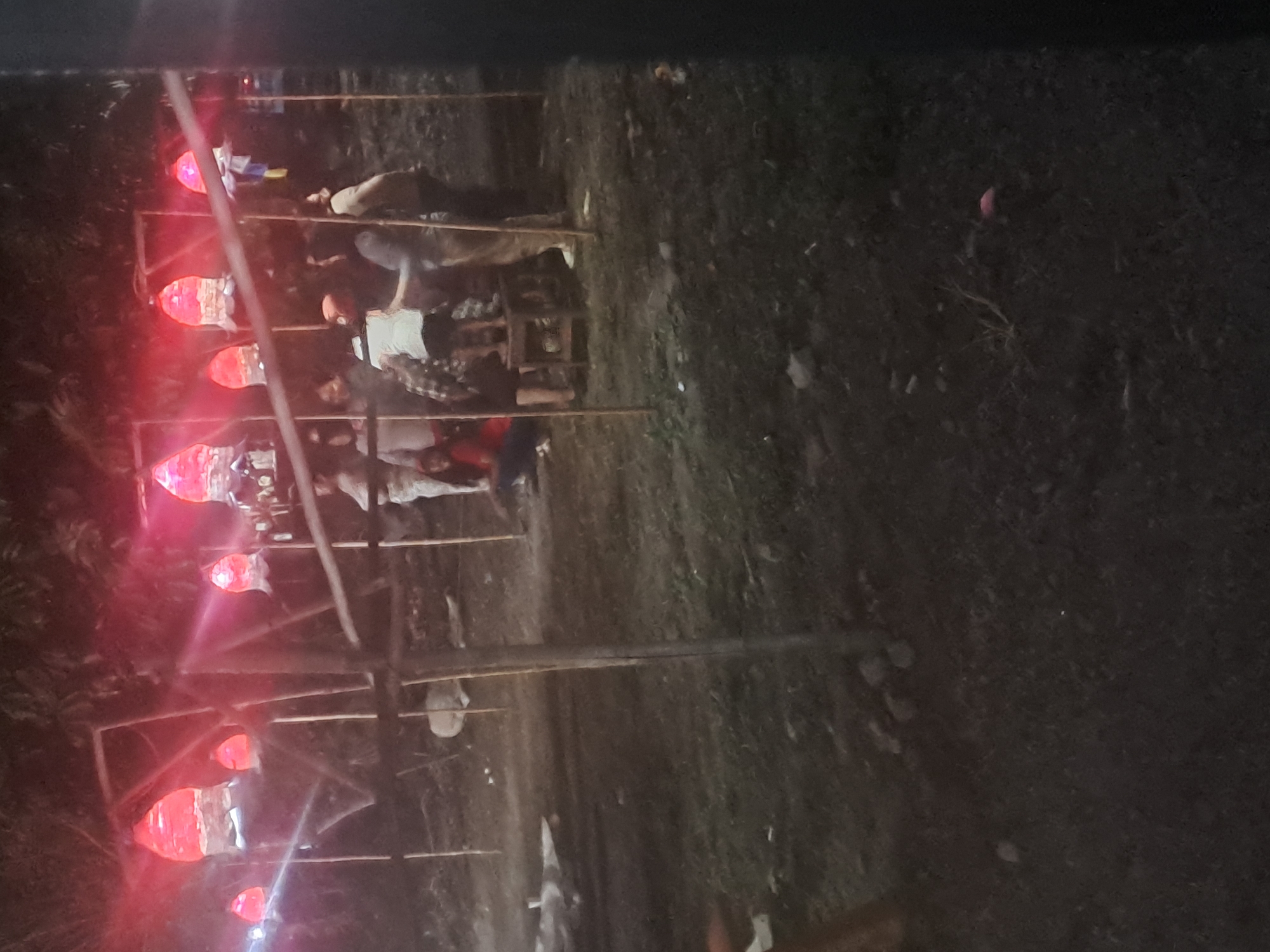 The image shows a nighttime outdoor scene with several illuminated, red, lantern-like structures suspended from wooden frames. A group of people are gathered beneath these lights. One person is standing on a small wooden stool. The ground is covered with grass and scattered debris. The overall impression is of a public gathering or festival in Tirtomartani, Indonesia.