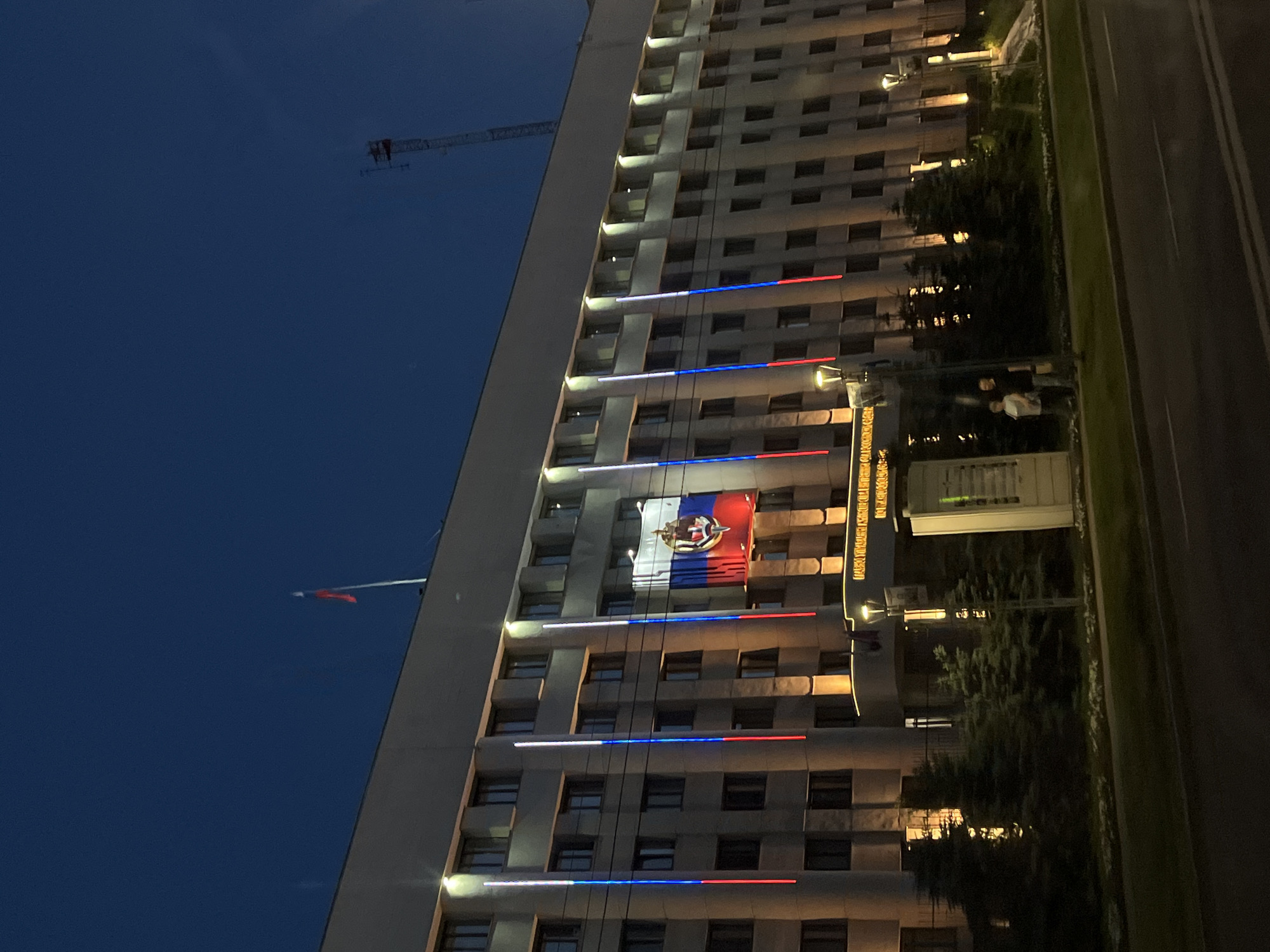 The image shows the building of the Nizhny Novgorod City Duma at dusk. The building, which is several stories high, is illuminated with warm lighting, highlighting its windows. Vertical light columns in the colors of the Russian flag (white, blue, and red) are displayed on the facade. A large banner with the Russian flag and coat of arms is also prominently displayed. In front of the building, there are several evergreen trees. A street with cars is visible in the foreground, and two individuals are standing on the sidewalk near the building. A radio tower is visible in the sky in the upper right portion of the image.