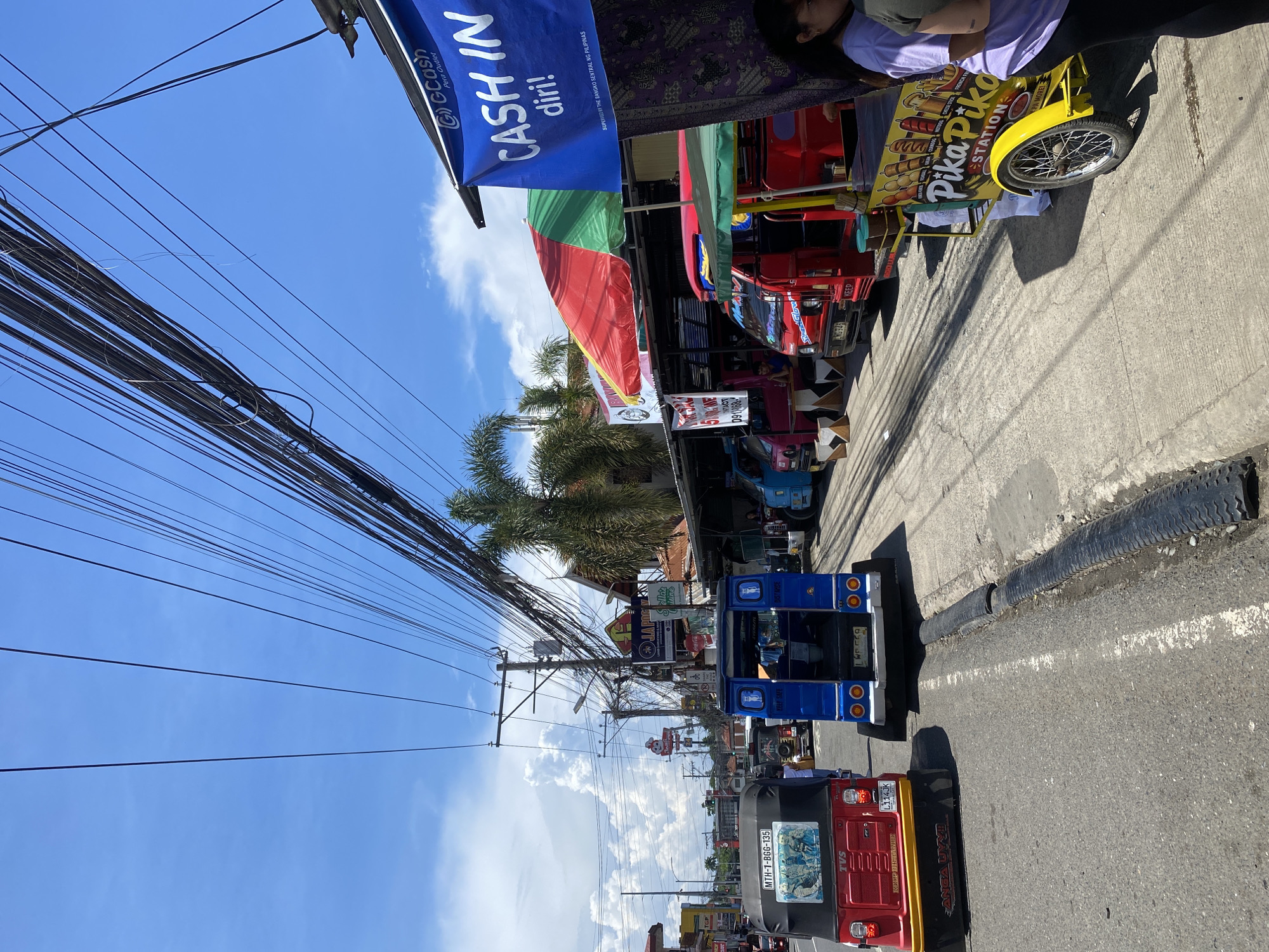 The image shows a street scene in Davao City, Philippines, under a clear blue sky with scattered clouds. Several vehicles, including a red and yellow tricycle with "TVS" branding and "MTH-1-BGG-135" license plate, and a blue jeepney with "KEEP SAFE" and "BANTAY" written on it, are on the asphalt road. The road has a raised speed bump covered with black material.
Along the side of the road, there is a row of stalls and shops. A blue banner that reads "GCash CASH IN diri!" is visible, indicating a financial service outlet. Several colorful umbrellas are set up over some of the stalls. One stall, painted yellow, has a sign that says "Pika Pika STATION" and displays images of food items.
The environment appears to be a busy urban area with many overhead electrical wires creating a complex network against the sky. Palm trees are visible in the background, suggesting a tropical climate. People are seen at some of the stalls, including a woman wearing a purple t-shirt and black leggings. The overall impression is that of a bustling street market or commercial area in a Philippine city.