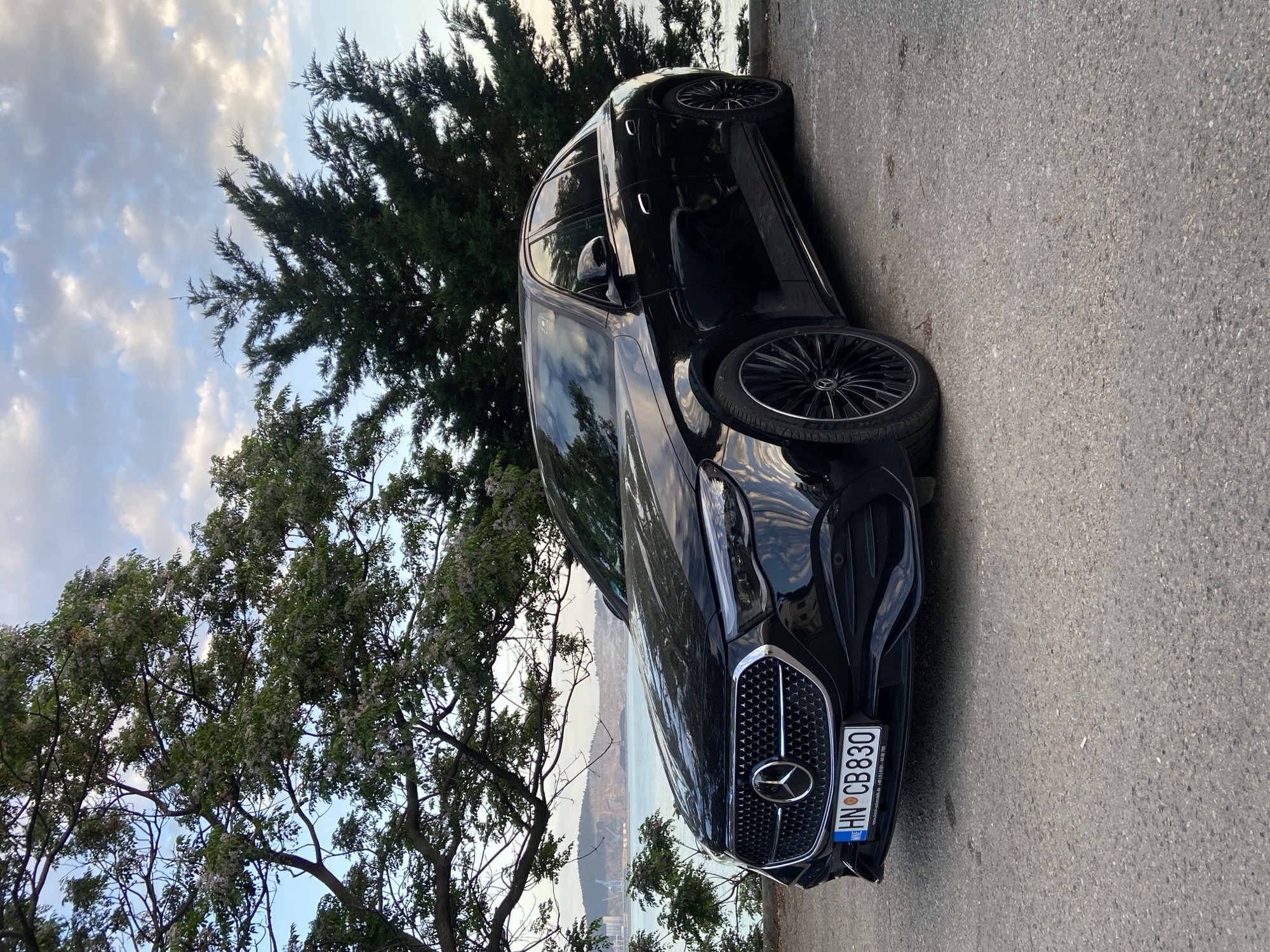 A black Mercedes-Benz is parked on the side of a road, with trees surrounding the car. The license plate reads "HN CB 830". In the background, there is a view of the sea and city of Bar, Montenegro. The sky is visible through the trees, with clouds.