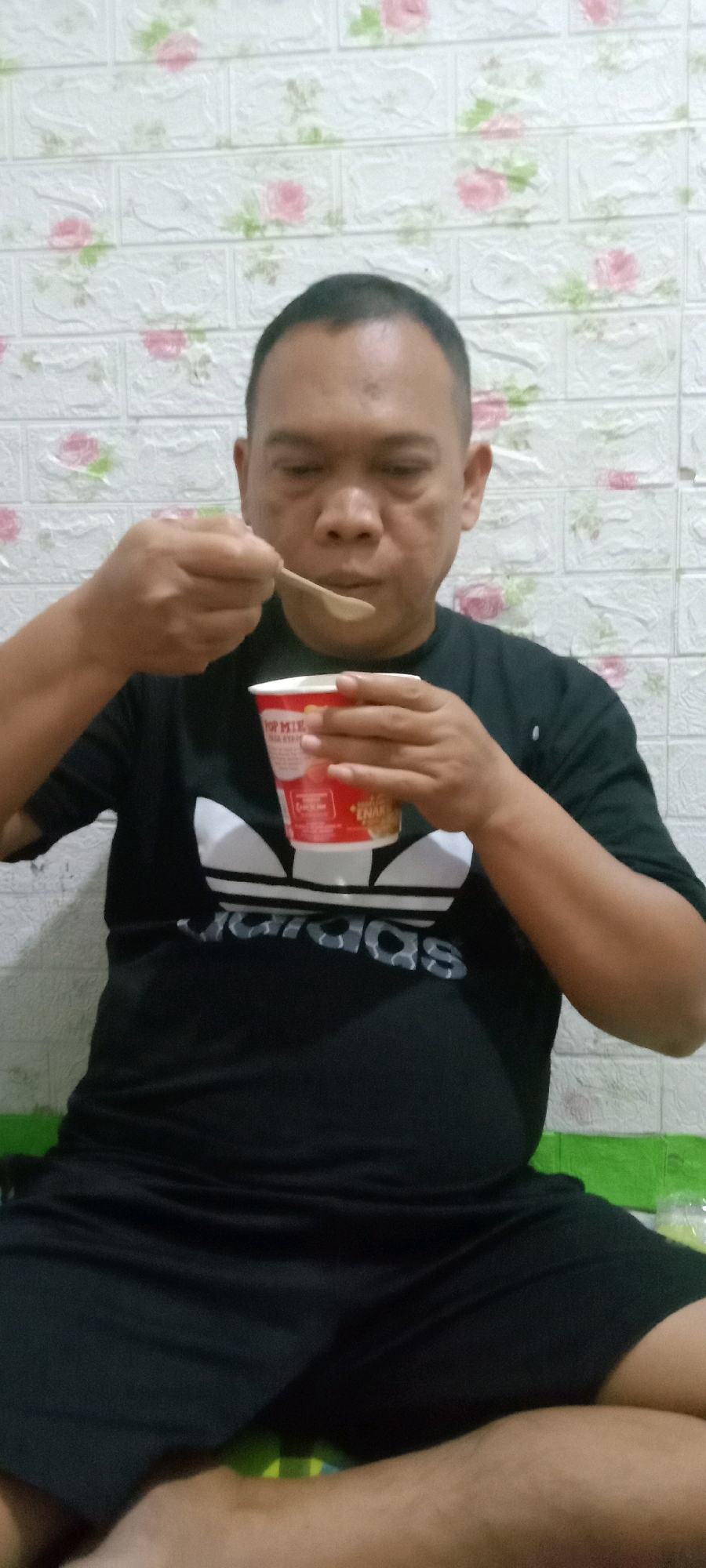 A middle-aged Indonesian man with a shaved head is eating from a red and white disposable cup. He is wearing a black Adidas t-shirt and black shorts. He is holding a wooden spoon in his right hand and the cup in his left hand. The background is a white brick-patterned wallpaper with pink and green floral accents. The man appears to be indoors, possibly in a home setting in Tasikmalaya, Indonesia.
