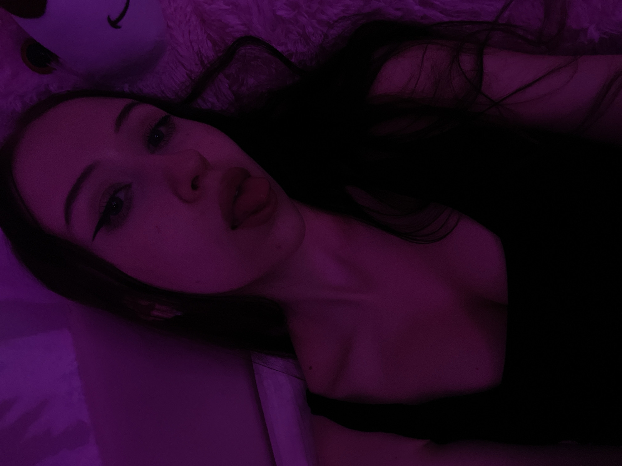 This image is a close-up selfie of a young woman in a dimly lit room, with purple ambient lighting. She has dark hair, prominent winged eyeliner, and her tongue is sticking out slightly. She is wearing a black tank top. A plush, light-colored object, possibly a stuffed animal, is visible in the background to her right. The overall impression is of a casual, private moment. The image does not contain any information about the location being Votkinsk, Russia, or any other specific geographical location.