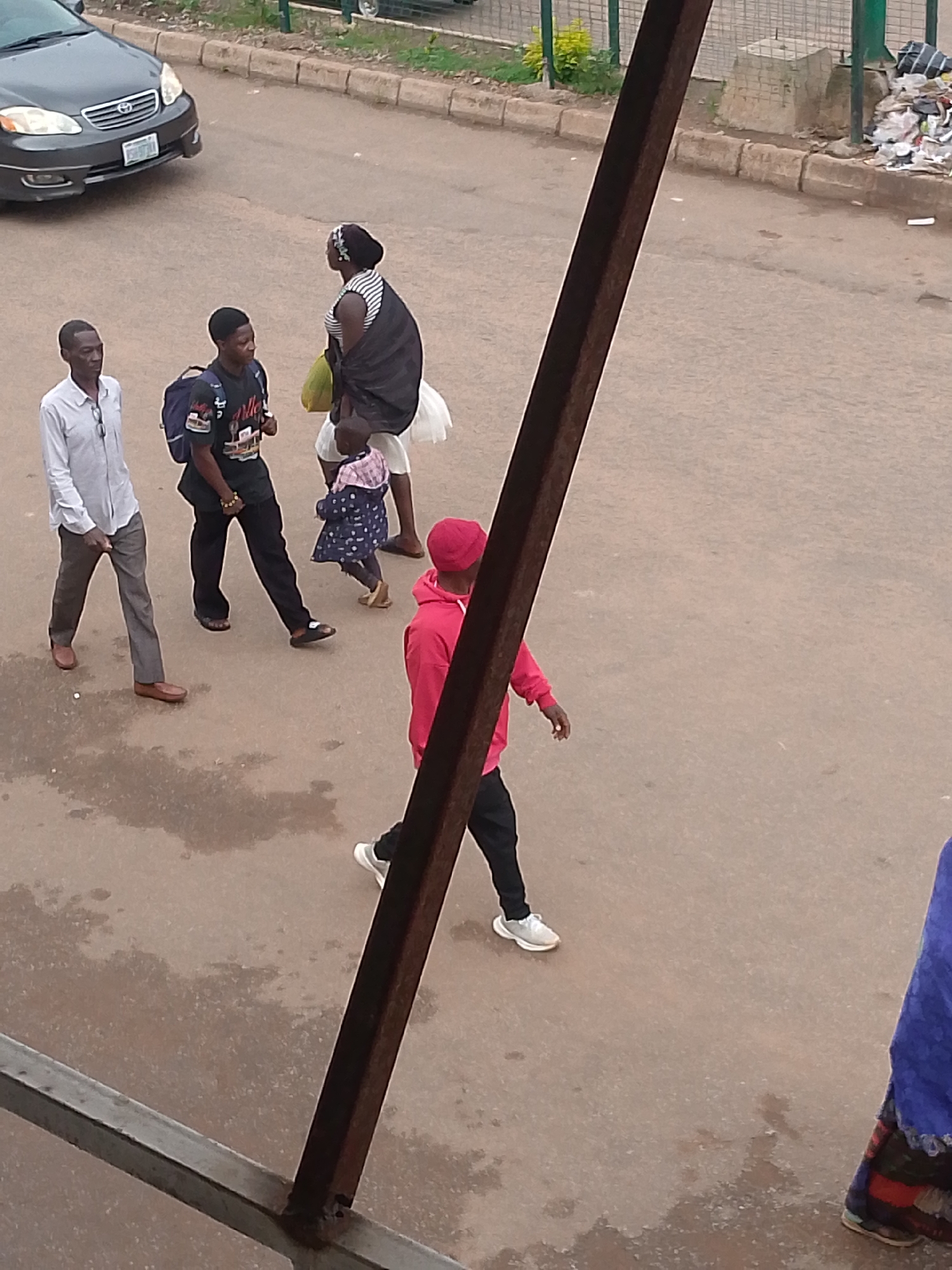 A group of people are walking on a paved street in Bukuru, Nigeria. The street is wet in patches, suggesting recent rain or a sprinkler. In the background, there is a curb with vegetation growing on it and a pile of trash. A dark grey car is parked on the left side of the image.
In the foreground, a man in a light blue collared shirt and grey pants walks to the left. Next to him, a man in a black t-shirt and black pants with a backpack walks in the same direction. Further to the right, a woman in a striped top and a black cloth draped over her shoulders walks with a young child in a blue polka-dot dress. A person in a bright pink hoodie and black pants walks away from the camera on the right side of the frame. Part of a blue and red patterned fabric is visible at the very bottom right of the image. A dark, rusty metal beam runs diagonally through the center of the image.