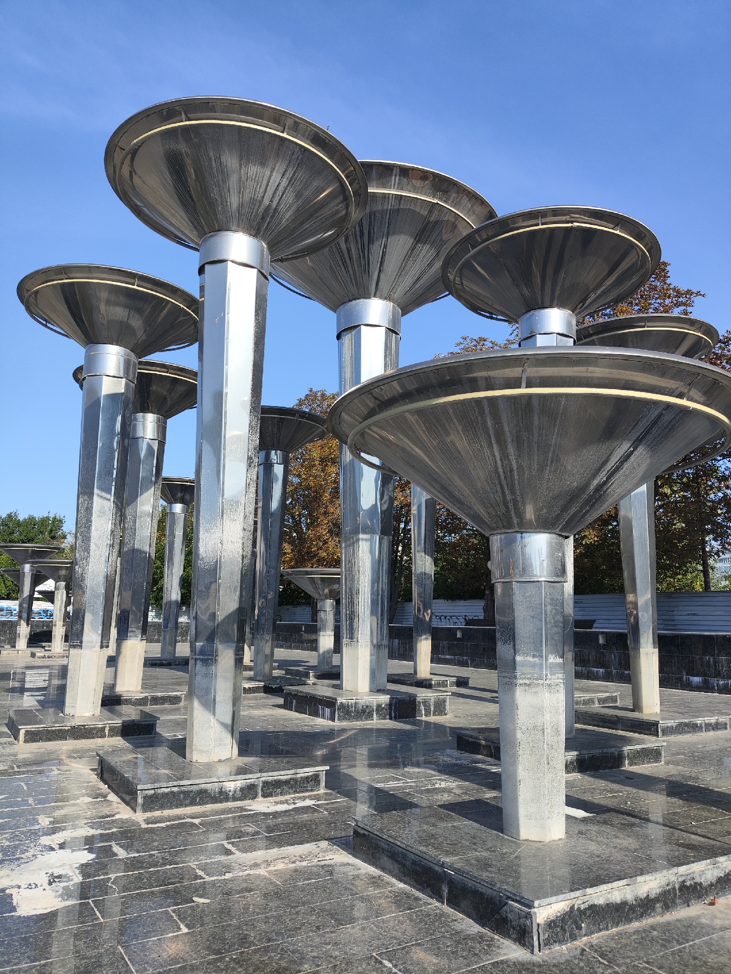 The image depicts a public plaza in Kharkiv, Ukraine, dominated by a collection of modern, metallic sculptures resembling inverted funnels atop tall, hexagonal pillars. These sculptures are arranged in a staggered formation across a dark, reflective tiled surface. The sky above is a clear, bright blue. In the background, trees with autumnal foliage and a fence are visible, suggesting the season is likely autumn. The overall impression is one of an outdoor public space, possibly a monument or a decorative element within a park or city square.
