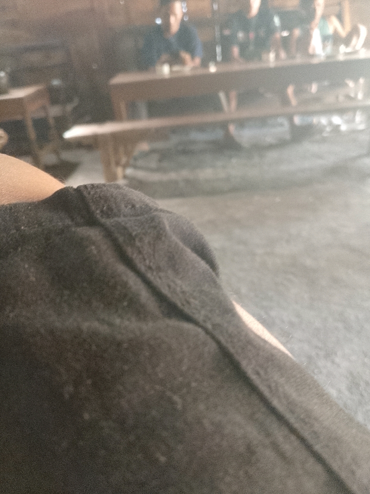 A close-up of a black garment in the foreground is out of focus, obscuring most of the background which appears to be a dimly lit indoor space. The background, though blurry, suggests a casual dining or gathering area with wooden tables and benches. Several individuals are partially visible seated at the tables, their faces indistinct. The overall impression is of a photograph taken in a dimly lit, possibly rural, Indonesian setting, perhaps a warung or simple eatery in Wadang.