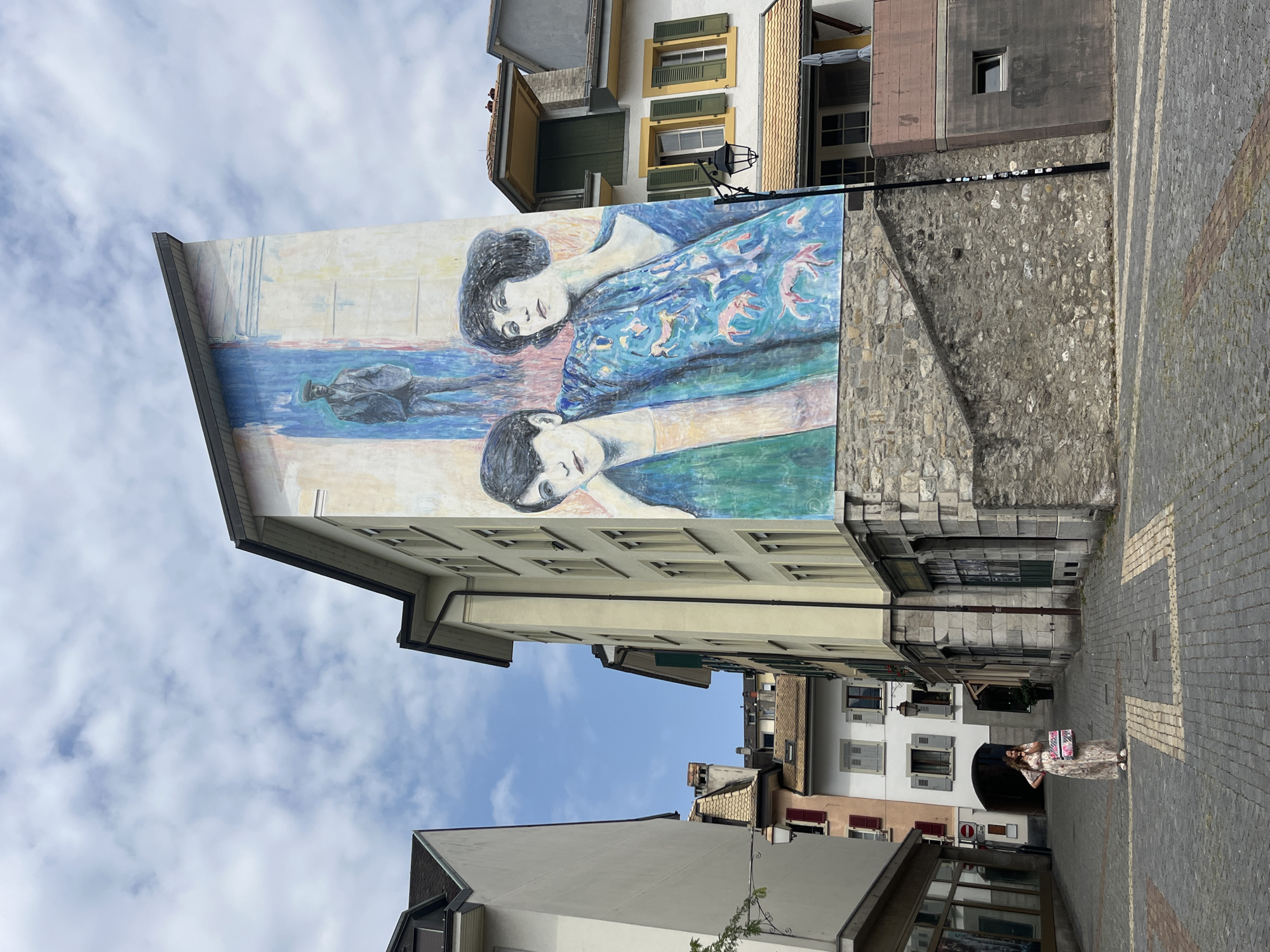 This is a photograph of a mural painted on the side of a building in Vevey, Switzerland. The mural depicts two women and a man. The women are in the foreground, and the man is in the background. The women are dressed in period clothing, and the man is wearing a suit. The mural is brightly colored, and the women are depicted in a realistic style. The building itself is tall and narrow, and it is located in a city alley. The alley is paved with cobblestones, and there are shops on either side of the street. A woman in a long floral dress is standing in the middle of the alley, holding a patterned bag. The sky above is cloudy.
