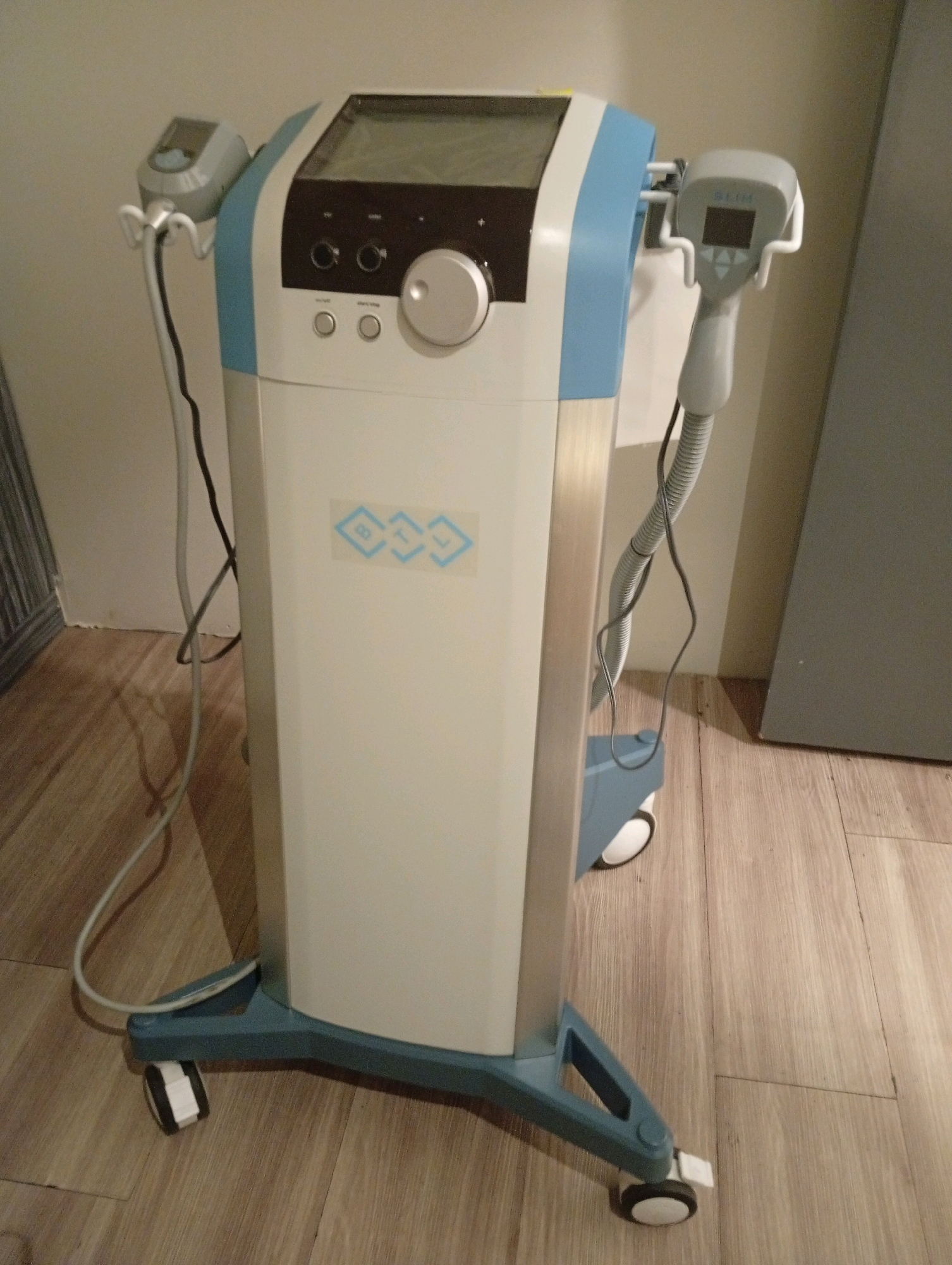 A medical device, likely for cosmetic treatments such as fat reduction or body sculpting, stands upright on a wooden floor. The device is predominantly white and light blue, with a silver metallic section. It features a control panel with a screen, buttons, and a large dial. Two hand-held applicators are attached to the sides of the device via flexible hoses, one with a smaller screen displaying "SLIM." The device is mounted on a blue wheeled base. The background consists of a light-colored wall and a gray door. The setting appears to be an indoor treatment room within a clinic or spa, possibly located in the city of Selayang Municipal Council, Malaysia, though no specific local landmarks are visible. There are no people present in the image.