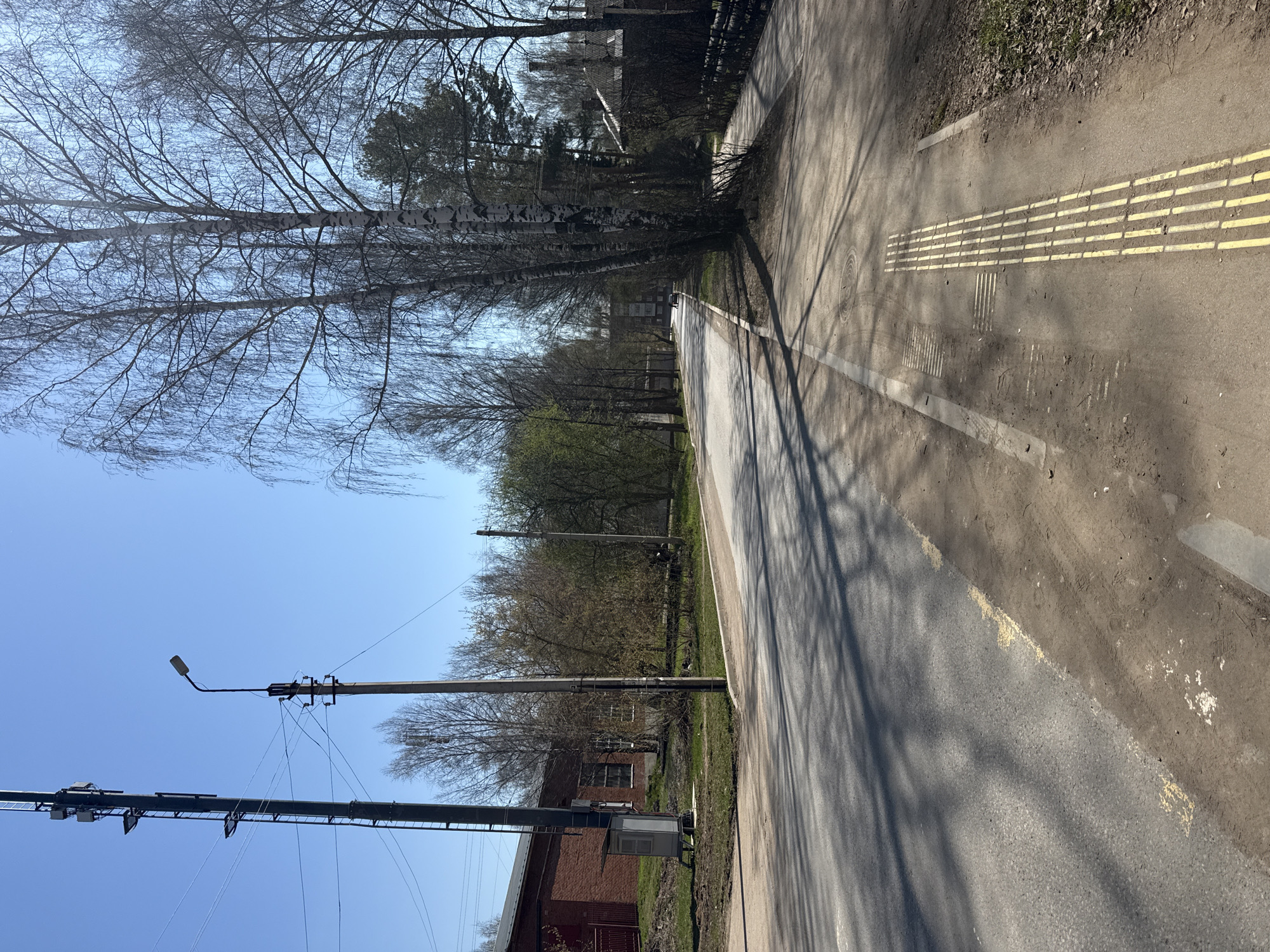The image shows an asphalt path in Votkinsk, Russia. The path has tactile paving markers for the visually impaired. A line painted on the path is partially faded. To the left of the path are trees, utility poles, and a brick building. The sky is blue and clear. The perspective is an eye-level shot taken at an angle. Shadows from the trees are cast onto the path. The trees are mostly bare, suggesting early spring.
