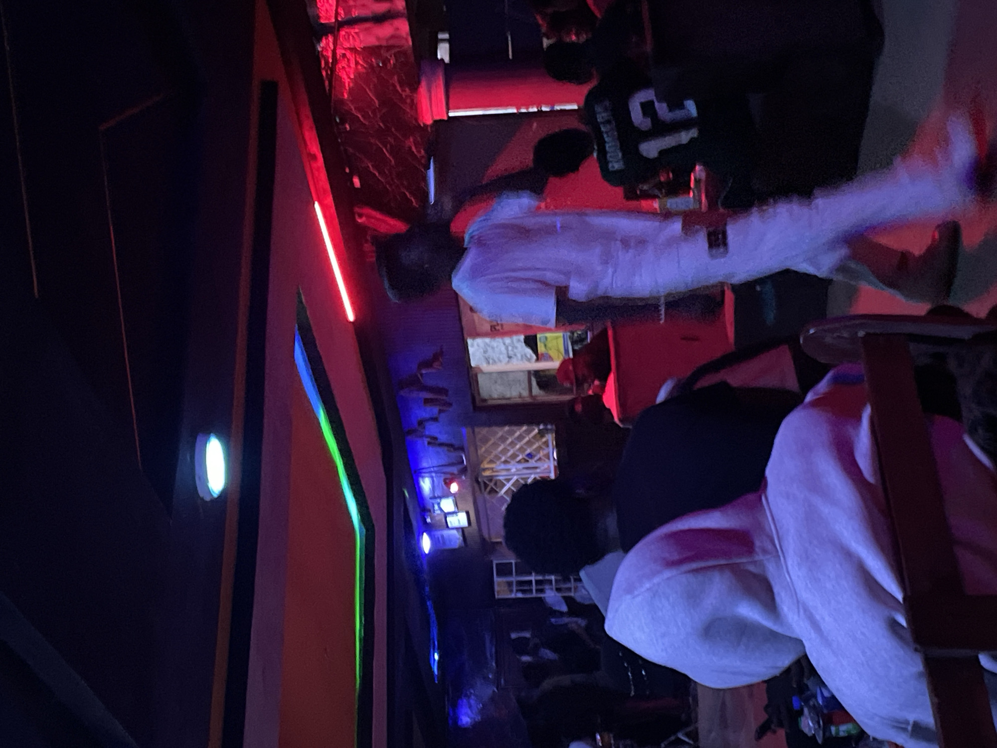 The image depicts an indoor scene, possibly a nightclub or event venue, with vibrant and colorful lighting. Dominating the foreground is a person wearing a white t-shirt and white pants, appearing to be in motion, possibly dancing or performing. To the left, two individuals are visible from behind, one wearing a light grey hoodie and the other a dark shirt. In the background, other individuals are present, some seated, and a person wearing a dark jersey with the number "12" and the name "RODGERS" is also visible. The environment features a stage-like area with neon lighting in green, blue, and red hues accentuating the ceiling and walls. The overall atmosphere suggests a lively gathering. The image does not provide enough information to definitively place the location in Durumi, Nigeria.