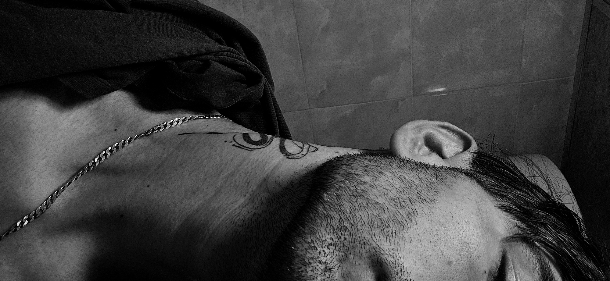 The black and white image depicts a person lying down. The focus is on the person's upper chest, neck, and head. A silver chain is visible around the neck. There is a tattoo on the shoulder. A dark-colored piece of clothing is draped over the shoulder. The person's face is partially visible, showing some hair and a portion of the eye and eyebrow. The background features tiled walls. There is no indication of the location being Kostenets, Bulgaria.
