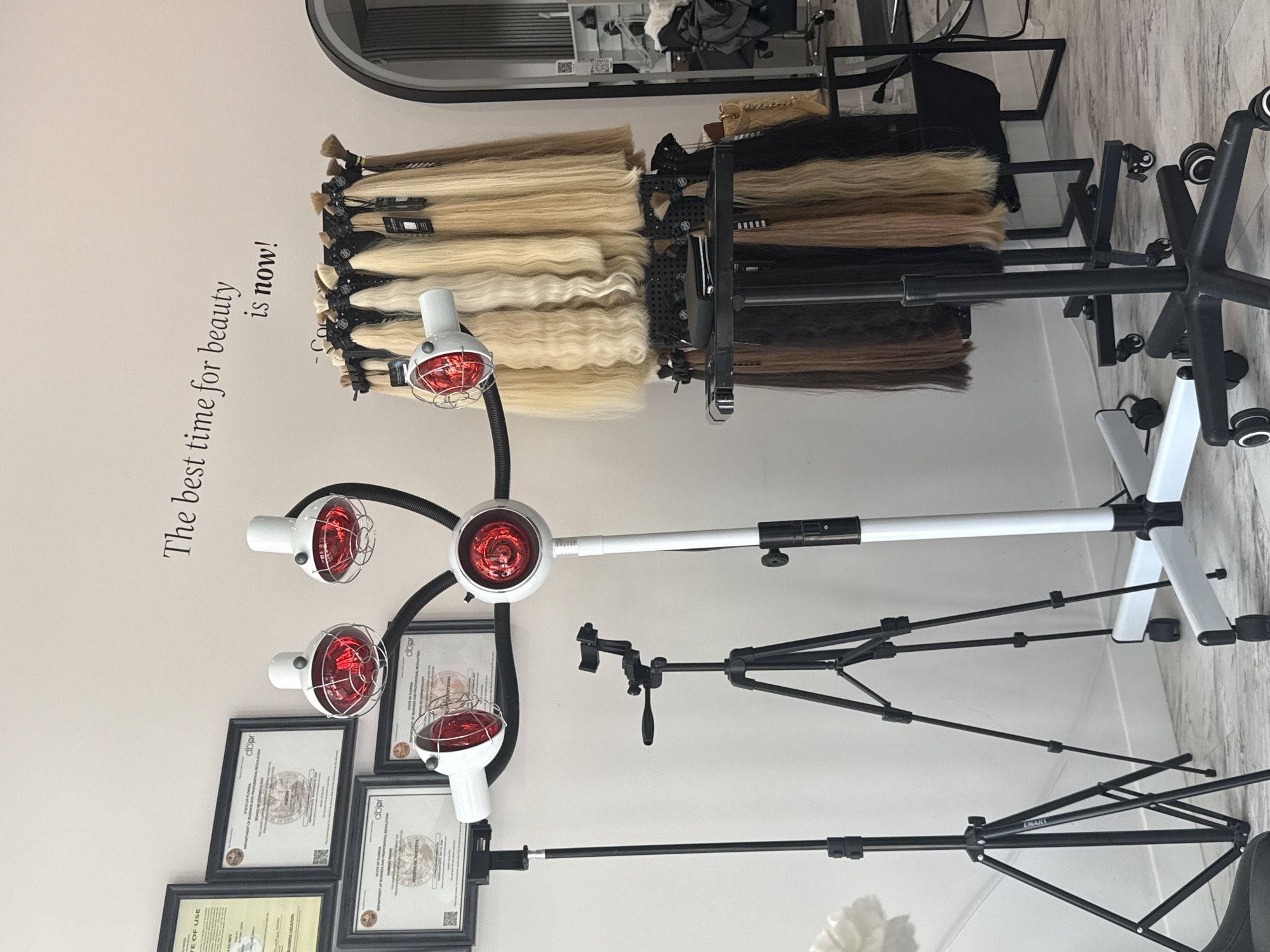 This image captures an interior view of what appears to be a beauty salon or hair extension studio in Hollywood, United States of America. A prominent display of human hair extensions is arranged on a rack, showcasing a variety of colors including different shades of blonde, brown, and black. Some of the blonde extensions exhibit wavy textures.
In the center of the frame stands a white, multi-armed device featuring several red-light lamps, likely infrared heaters used for hair treatments, processing, or drying. To the right of this, two black tripod stands are visible, one equipped with a phone or camera holder. A black office chair with wheels is partially seen in the upper right background.
The wall on the left side of the image features framed certificates, which include discernible text such as "STATE OF FLORIDA" and "dbpr" (Department of Business and Professional Regulation), suggesting professional licensing or accreditation. One of the certificates and one of the tripods also bear the text "EMARI". Above these frames, a motivational quote is stenciled on the wall: "The best time for beauty is now!". The flooring has a light-colored, wood-grain appearance. The scene depicts a professional setting focused on hair services.