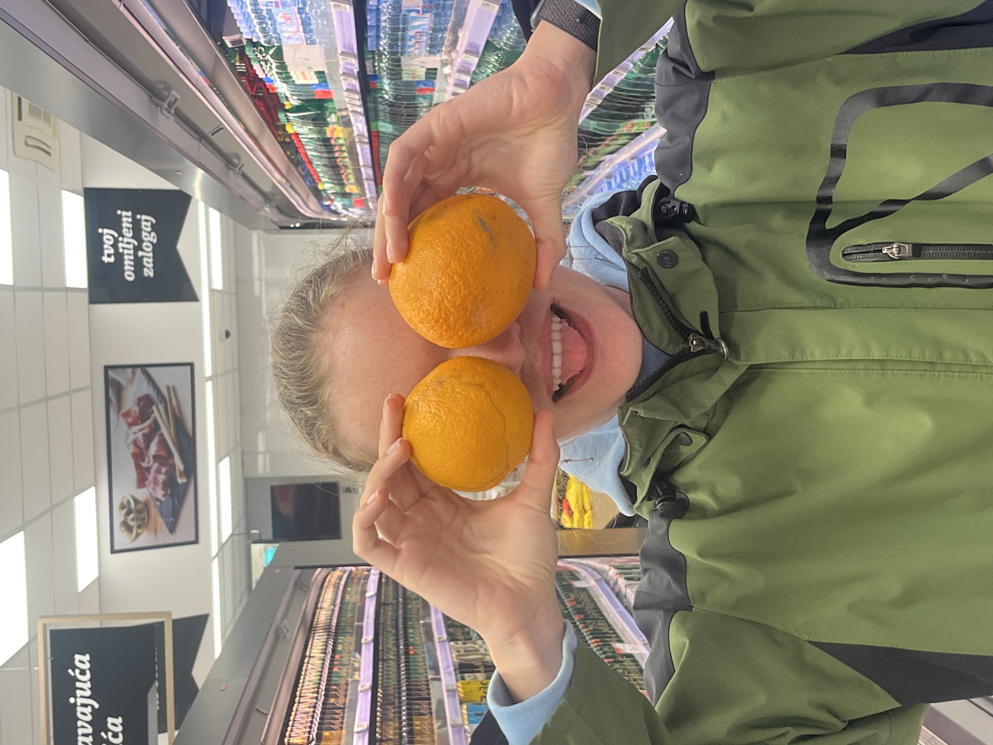 A person in a green and black jacket is holding two oranges in front of their eyes and mouth, appearing to be smiling. The scene is inside a grocery store, with shelves stocked with products visible in the background. Signs in a foreign language are also visible.