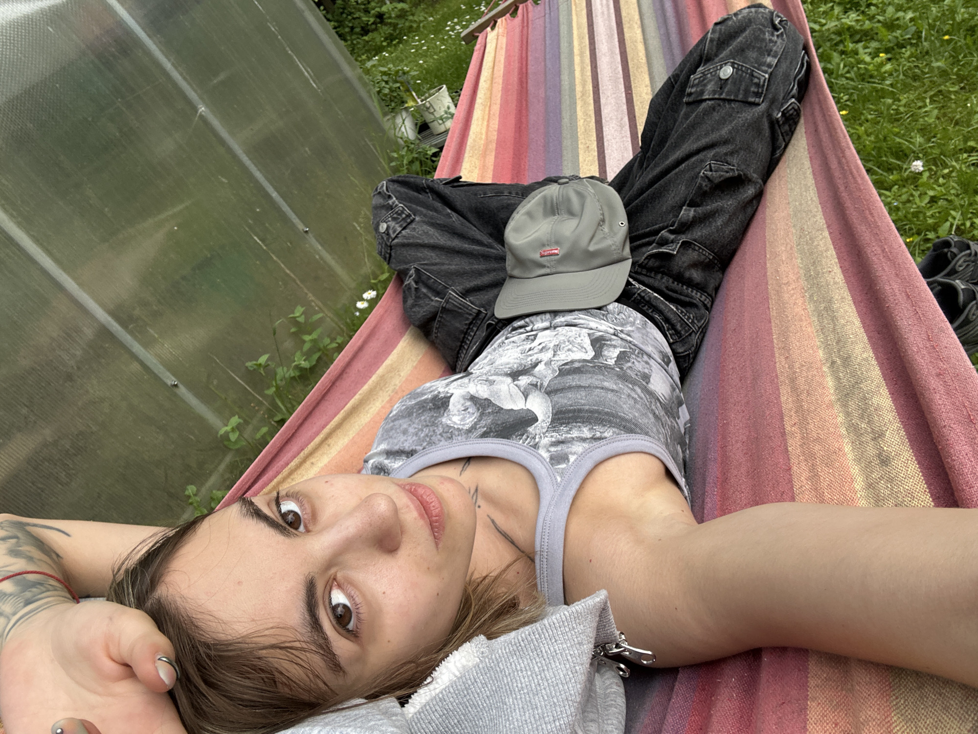 A person is lying in a hammock in an outdoor setting, possibly a backyard. The hammock has colorful stripes in shades of red, yellow, and pink. The person is wearing a gray tank top with a graphic print, dark wash cargo pants, and a gray baseball cap with a red logo. The cap is placed on their lap. Their left arm is visible, showing tattoos.
In the background, there is a greenhouse with a translucent, corrugated roof. Green vegetation, including grass and some plants, surrounds the area. A few white flowers are visible in the grass near the hammock. The overall lighting suggests it is daytime, with soft natural light, implying possibly an overcast or late afternoon setting.
The person's face is turned towards the camera, with eyes open and looking directly at it. Their expression appears relaxed and perhaps a little pensive. The image has a casual, informal feel.
There is visible text on the red logo on the gray baseball cap, which appears to be "Supreme".