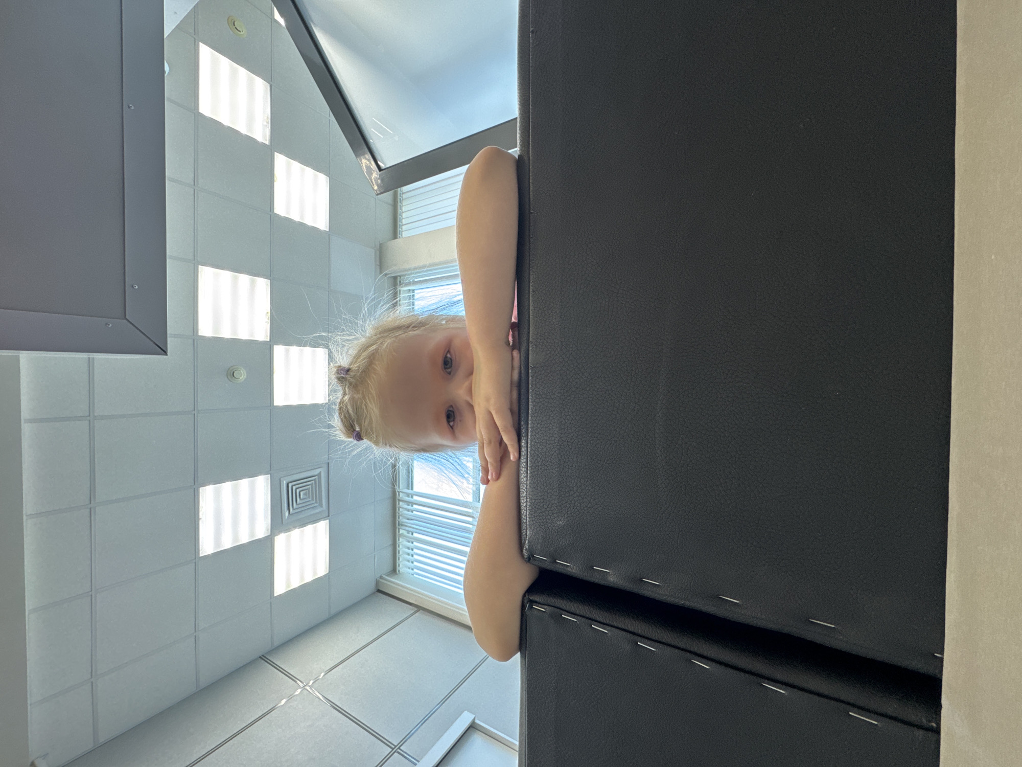 A child with blonde hair is peering around the edge of a dark, possibly black, rectangular object. The setting is indoors, indicated by the tiled walls and recessed lighting above. A window with blinds is visible. The city of Perm, Russia is not verifiable from this image.
