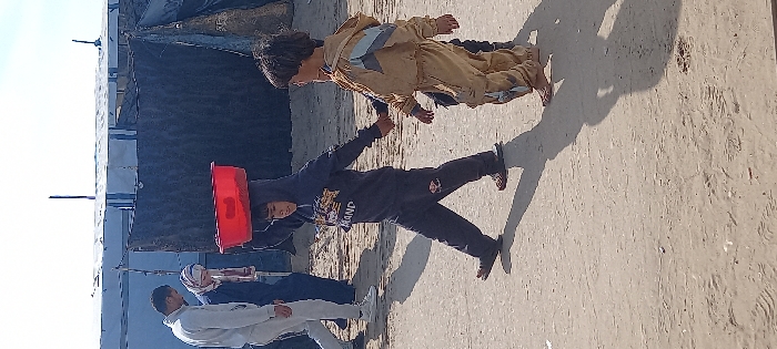 The image captures an unpaved, sandy area in Khan Yunis, Palestinian Territory, under a clear sky, featuring five individuals and makeshift structures. In the foreground, two young children walk from left to right, holding hands. The child on the left, wearing a dark hooded tracksuit with a light-colored graphic print and dark sandals, carries a red plastic basin balanced on their head, steadying it with their left hand. The child on the right, with dark hair, wears a light brown tracksuit featuring light blue or grey patterns on the upper back and appears barefoot. Behind the children, an adult male in a light grey tracksuit and white sneakers walks from left to right. Further back, partially obscured by the foreground subjects, an adult wearing a dark garment and a light head covering is visible, along with another individual in a patterned shirt. The background consists of several rudimentary structures constructed from blue tarpaulins, dark fabric, and sheeting, with poles extending upwards, consistent with an informal settlement.