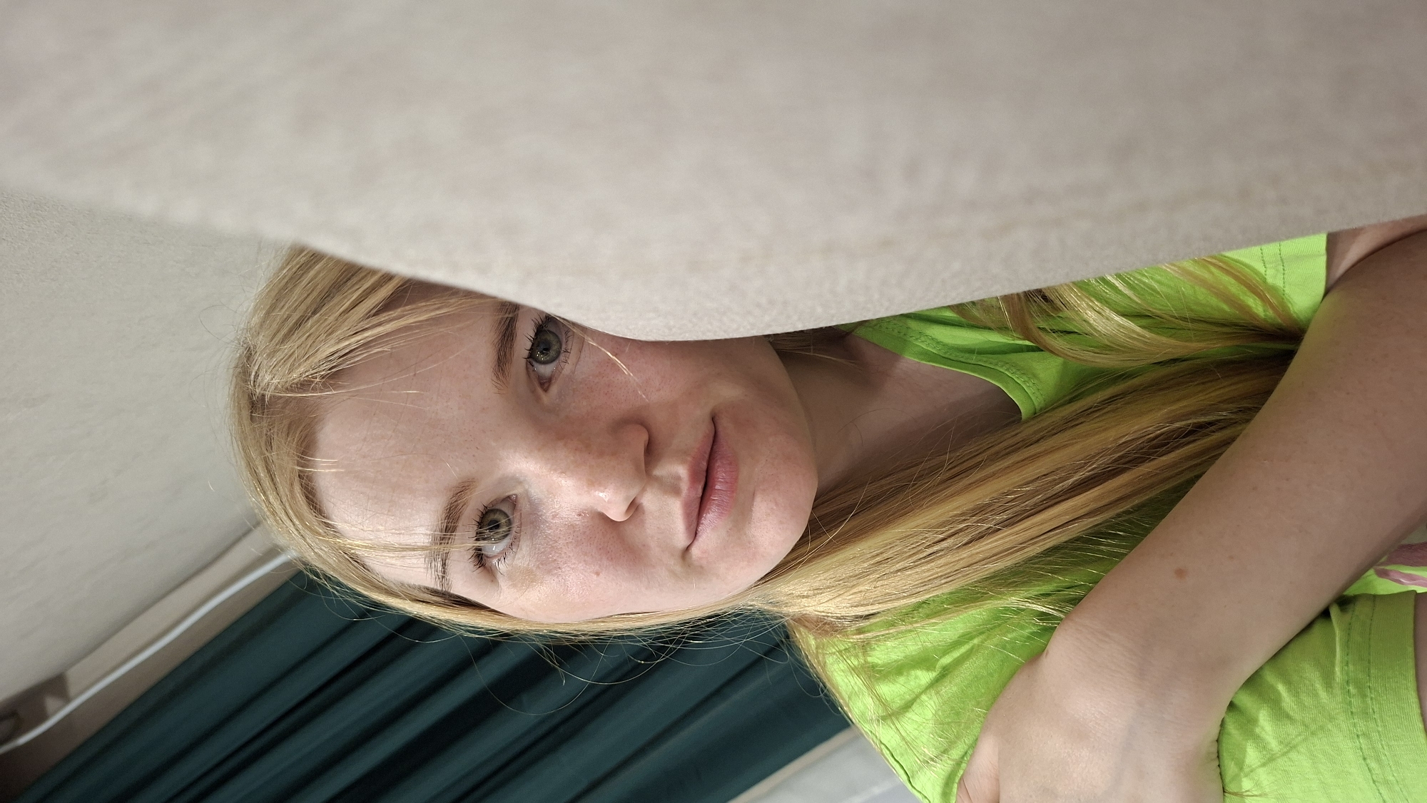The image features a close-up, upside-down shot of a young woman with blonde hair and fair skin with freckles. She is wearing a bright green t-shirt. Her eyes are a light green-hazel color, and her lips are slightly parted. She appears to be lying down, possibly on a bed or couch, as a light-colored textured surface is visible above her head, obscuring part of her face and hair. The background shows dark green drapes, suggesting an indoor setting, possibly a bedroom or living room. The lighting is soft, and there are no obvious indicators of the time of day or weather conditions. The woman's expression is relaxed, with a slight smile. There is no visible text in the image. The overall impression is an intimate and candid moment captured from an unusual perspective.