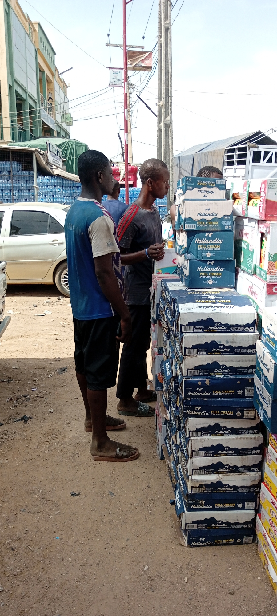 Two dark-skinned men stand beside a large stack of boxed Hollandia Full Cream Evaporated Milk, a Chivita juice box, and bottled water. The men are dressed casually in t-shirts, shorts, and open-toed sandals. The environment suggests an outdoor market or storefront in Fagge A, Nigeria, based on the available information. In the background, there is a building, parked cars, and utility poles, adding context to the urban setting.