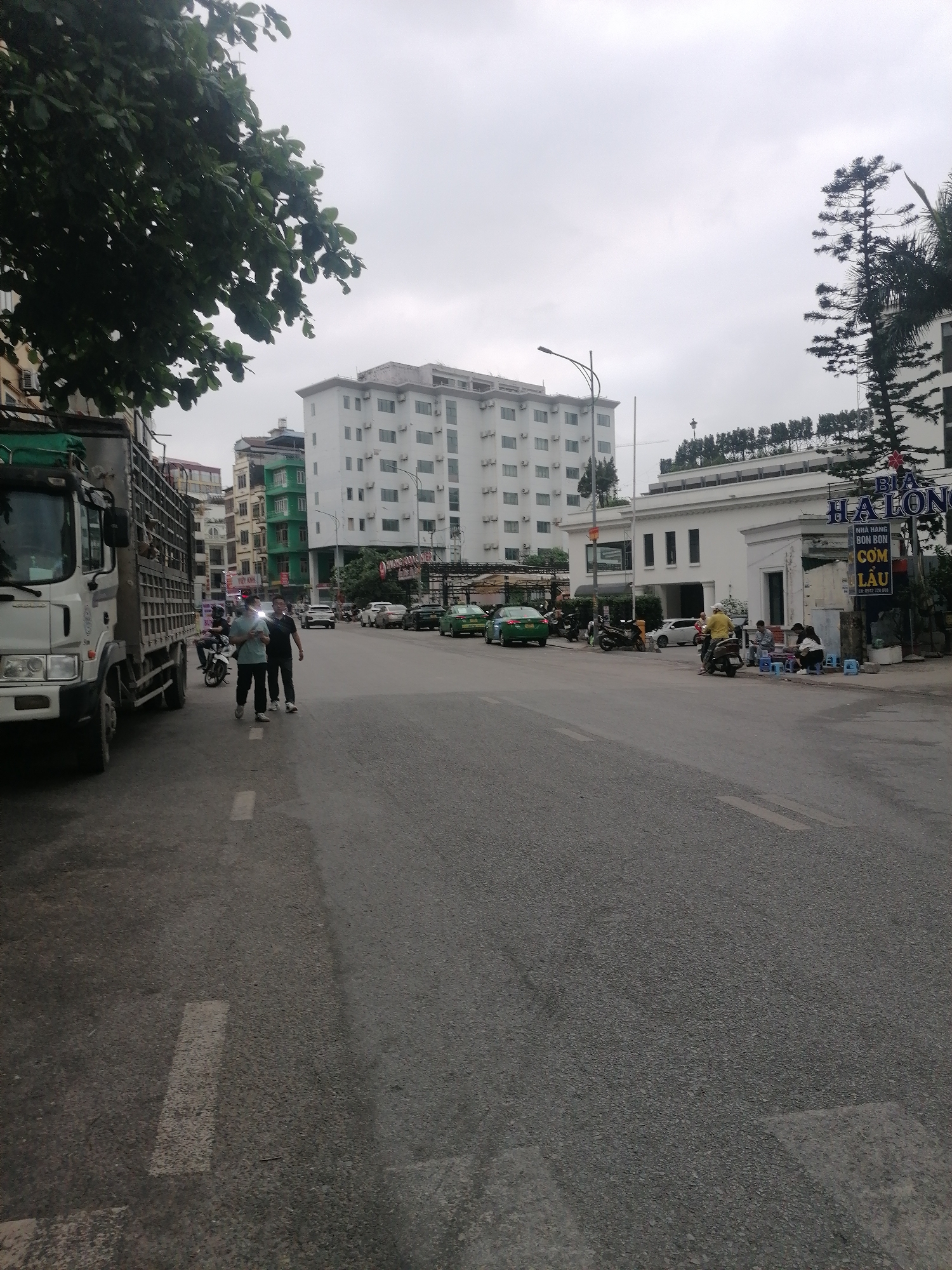 The image depicts a street scene in Bai Chay Ward, Vietnam, on an overcast day. A large white truck is parked on the left side of the road. Two men are walking across the street in front of the truck. Several green taxis and other vehicles are parked along the side of the street. People are seated on stools at a roadside eatery with a "BIA HA LONG" sign. A white building with a flat roof is visible behind the eatery. Taller buildings and trees are in the background. The road is asphalt and has painted lane markings.