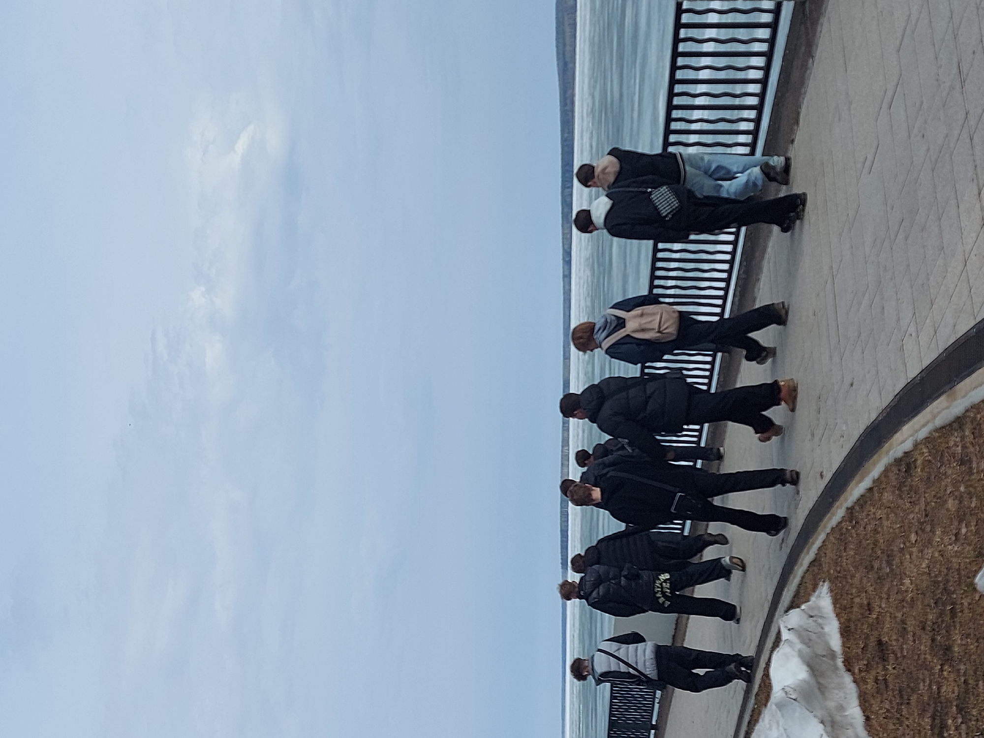 A photograph depicts a group of people standing along a promenade in Chaikovsky, Russia. They are looking over a railing towards a body of water, possibly a frozen river or lake, beneath a cloudy sky. The promenade is paved and has a decorative border, with some snow patches. The people are dressed in casual winter clothing.