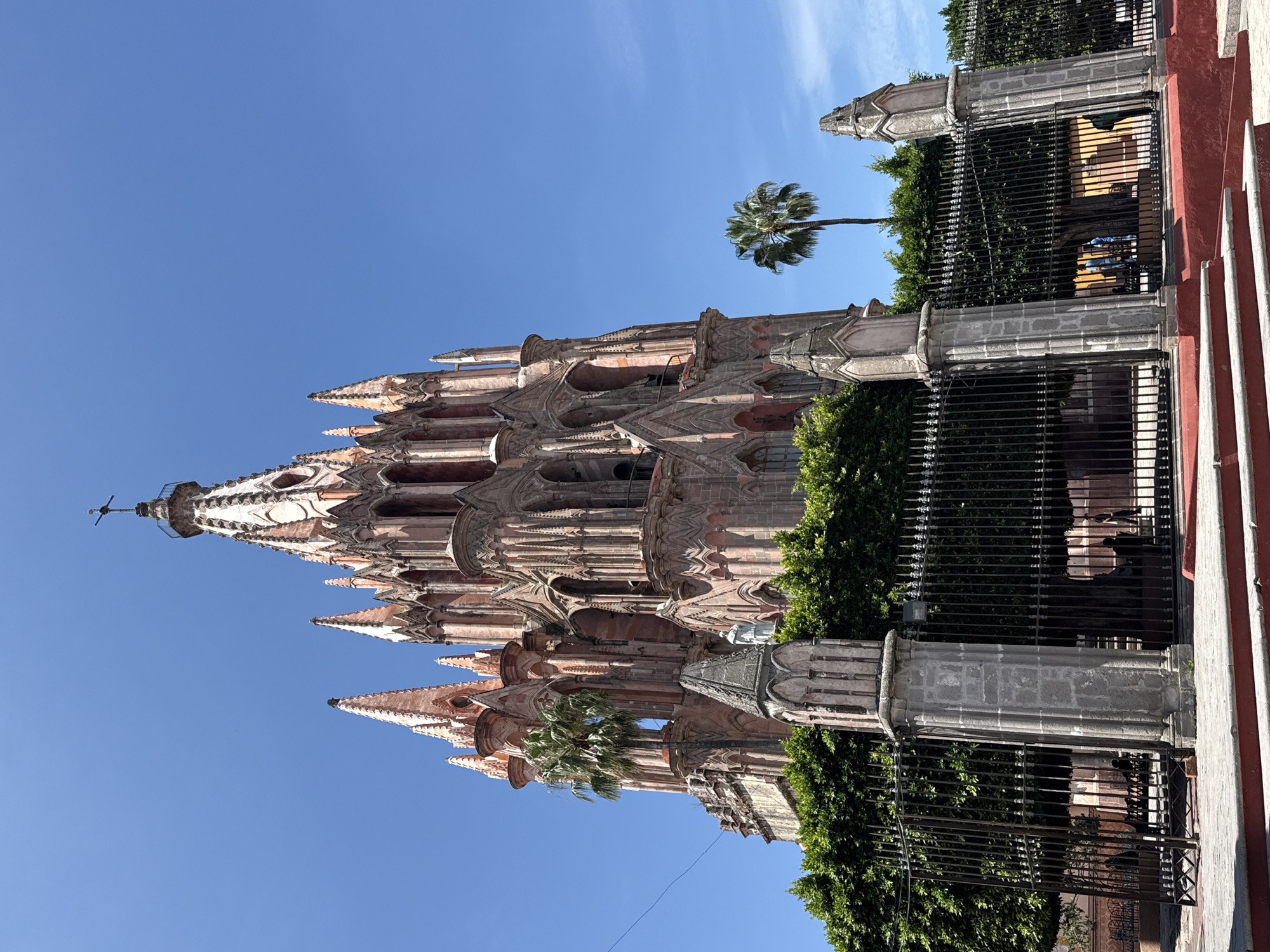 The image shows the iconic Parroquia de San Miguel Arcángel in San Miguel de Allende, Mexico, on a clear, sunny day. The neo-Gothic church, constructed from pink stone, dominates the frame with its tall, ornate spires topped with crosses. A large green tree and a tall, slender palm tree are visible to the left and right of the church, respectively. A black metal fence with stone pillars in front encloses part of the church grounds, with lush green foliage behind it. Red steps lead up to a sidewalk in the foreground. A few silhouetted figures are visible behind the fence, some appearing to be walking or standing. The sky is a vibrant, cloudless blue.