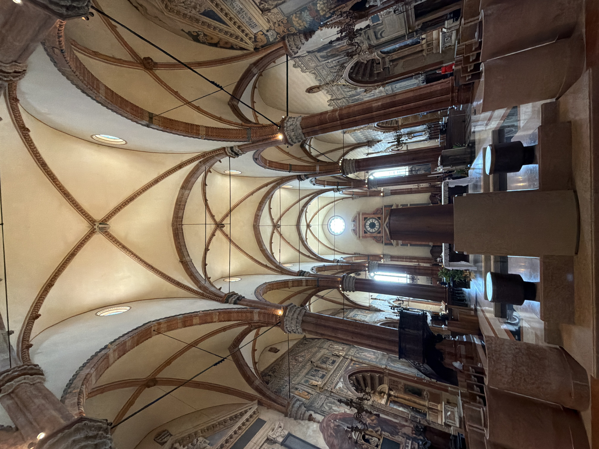 The image depicts the interior of the Basilica di Sant'Anastasia in Verona, Italy. The photograph captures the Gothic architecture of the nave, characterized by its high vaulted ceilings supported by red marble columns. The vaulted ceiling features intricate rib vaulting. Sunlight streams in from high windows, illuminating the space.
In the foreground, there are tiered stone structures and cylindrical wooden objects, possibly part of seating or decorative elements. Beyond these, the floor is paved with reflective tiles, showing a pattern of light and shadow.
The nave extends towards the altar area, where a white altar is visible. Surrounding the altar are pews, some empty and some with a few individuals seated. A large clock face is mounted on the wall in the distance, to the right of the central axis of the nave.
On the left side of the nave, a dark wooden pulpit is positioned on a column. The walls of the basilica are adorned with frescoes and architectural details, including arched niches and decorative friezes. Several chandeliers hang from the ceiling.
A few people are visible in the scene: one person in a bright red garment is standing near the right side aisle, and a few figures are seated in the pews towards the altar. The overall impression is a solemn and grand ecclesiastical interior.