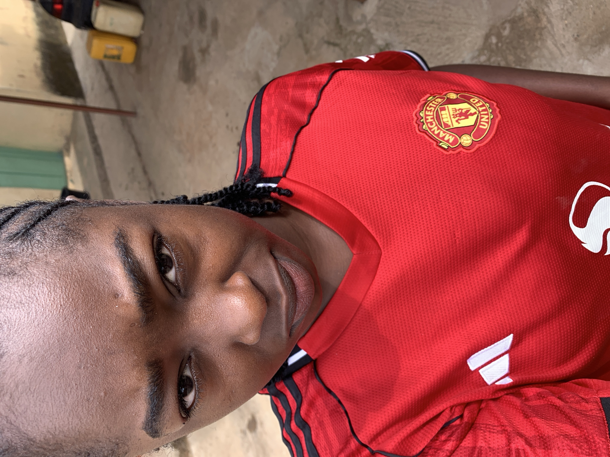 The image captures a close-up portrait of a person, likely female, with dark skin and braided hair, looking directly at the camera with a calm, slightly smiling expression. The subject is wearing a red Manchester United football jersey. The club's crest, featuring "MANCHESTER UNITED" text in yellow and red, is prominently displayed on the left chest. A white Adidas performance logo is partially visible on the right side of the jersey. The jersey material exhibits a textured pattern.
The background is subtly blurred, suggesting an outdoor or semi-outdoor setting in Rafin Par, Nigeria. A light-colored, possibly concrete or dirt, ground is visible, along with a neutral-toned wall or structure. In the upper background, two out-of-focus containers, one yellow and one lighter-colored, are discernible. The lighting indicates it is daytime.