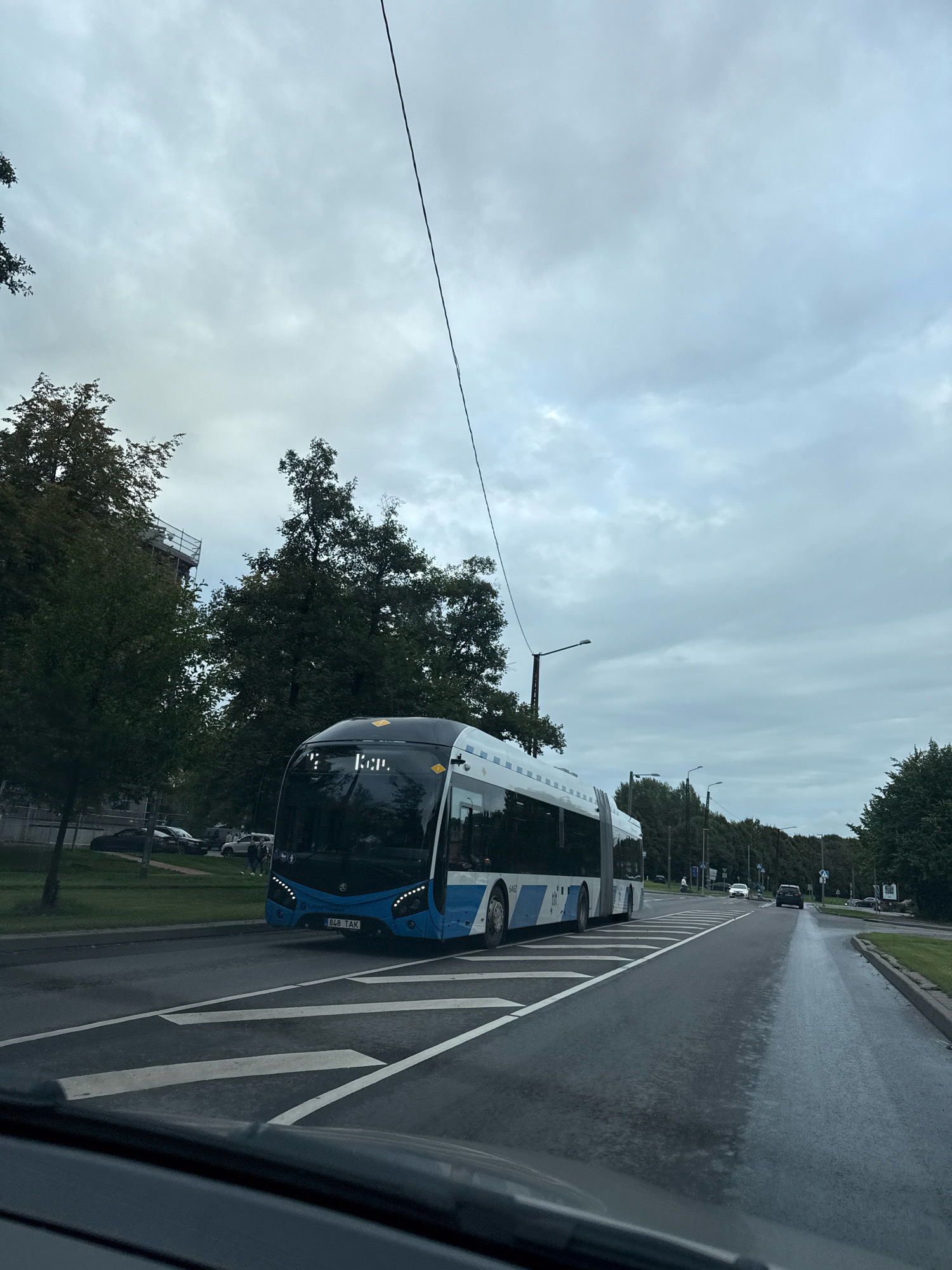 A blue and white articulated bus is driving on a wet asphalt road in Tallinn, Estonia. The bus has a license plate that reads "848 TAK". The sky is overcast with grey clouds. Trees line the side of the road, and there are streetlights and power lines visible. In the background, other vehicles and pedestrians can be seen. The image appears to be taken from inside another vehicle, possibly a car, looking out through the windshield.