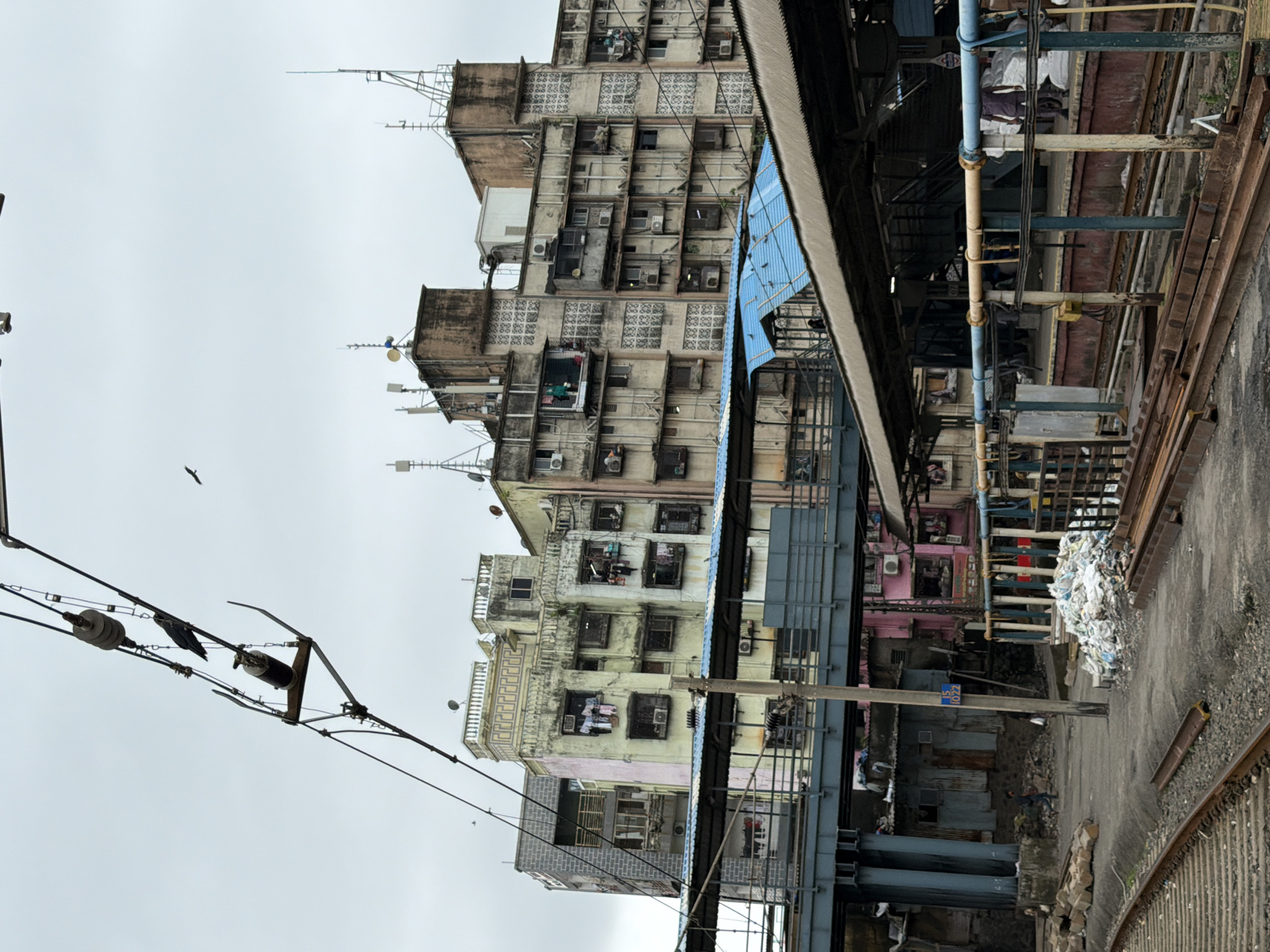 This is a vertical, eye-level, full shot of a railway station and surrounding buildings in Mumbai, India. The scene is cluttered with various elements, suggesting a densely populated urban environment. In the foreground, railway tracks are visible, with some rusted metal rails lying nearby. A concrete platform with a brown and red base runs parallel to the tracks. A covered walkway with a blue roof extends over the platform.
Beyond the platform, a raised pedestrian walkway, constructed with blue metal supports, crosses the frame. Behind this walkway, a series of tall, multi-story apartment buildings are visible. These buildings are closely packed together and appear old and somewhat dilapidated, with visible signs of wear and tear. Many windows are open, and clothes hang from some of them. Air conditioning units are mounted on the exteriors of several buildings. The rooftops are adorned with numerous antennas and satellite dishes. The sky is overcast and grey.
In the mid-ground, people are present. A person in dark clothing is walking along the platform. Another individual, wearing a blue shirt and dark pants, walks away from the viewer towards a pink-colored building structure. Near a wooden pole with a sign that reads "15 1022," a pile of white plastic trash bags is stacked. Debris and scattered building materials are also present on the ground. The scene is dominated by a sense of urban density and everyday life in a bustling Indian city.