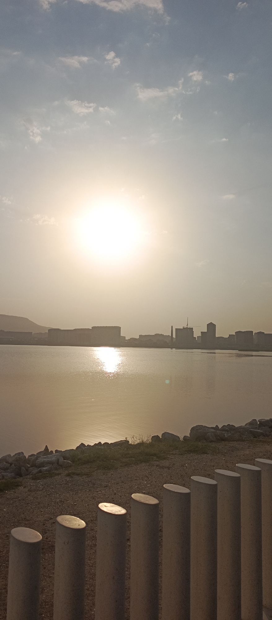 The image depicts a serene waterfront scene in Makhachkala, Russia, during what appears to be late afternoon or early evening. The sun, a bright, diffused orb, hangs low in the sky, casting a warm, golden glow across the water and the hazy skyline. The sun's reflection creates a shimmering path across the calm surface of the water.
In the foreground, a row of light-colored, cylindrical concrete bollards with angled tops are arranged in a line. Beyond them, a pebbled ground transitions into a rocky embankment bordering the water. Some sparse green grass and small plants grow amongst the rocks.
The middle ground is dominated by a body of water, likely the Caspian Sea, given the location. Across the water, a distant city skyline is visible, characterized by a collection of modern buildings, some appearing to be residential complexes, and industrial structures, including a prominent tower with a crane at its peak, suggesting ongoing construction. The buildings are silhouetted against the bright sky, softened by atmospheric haze. A hill or mountain range can be faintly seen in the far left of the skyline.
The sky above is a pale blue, transitioning to a lighter, almost white hue closer to the sun. Scattered white clouds drift across the sky, some catching the sunlight and appearing slightly luminous. The overall atmosphere is tranquil and expansive. No people are visible in the image.