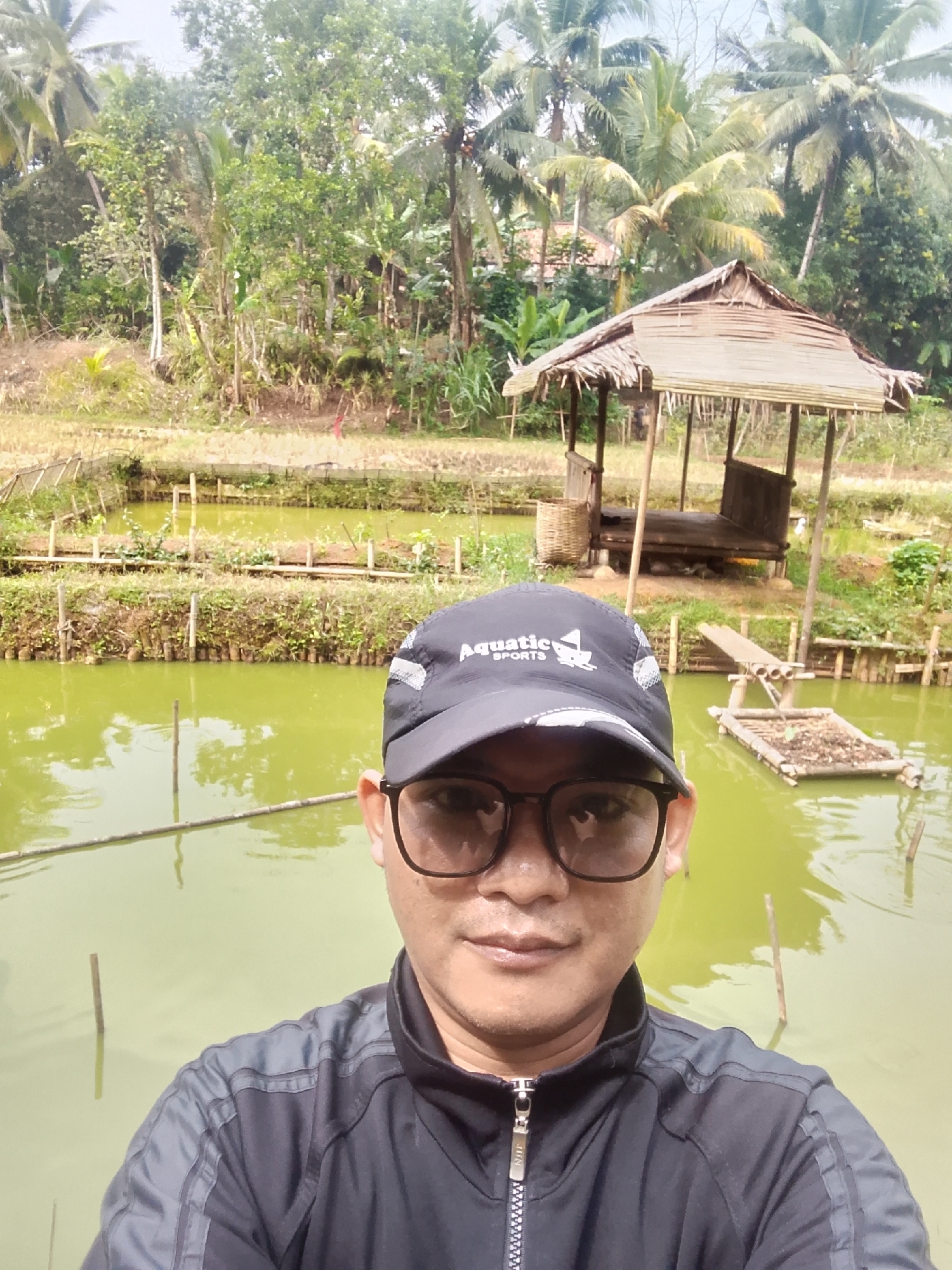 A selfie of a man wearing a black cap and jacket, posing in front of a natural landscape with ponds and lush vegetation in Cikukulu, Indonesia. The man's face is partially visible, wearing glasses. Behind him, there are several green ponds, some with wooden posts in the water. A rustic bamboo hut with a thatched roof is situated on the right side of the image, overlooking the ponds. The background is filled with dense trees and palm trees, indicating a tropical environment. The overall scene depicts a rural setting, likely a farm or a recreational area, surrounded by nature.