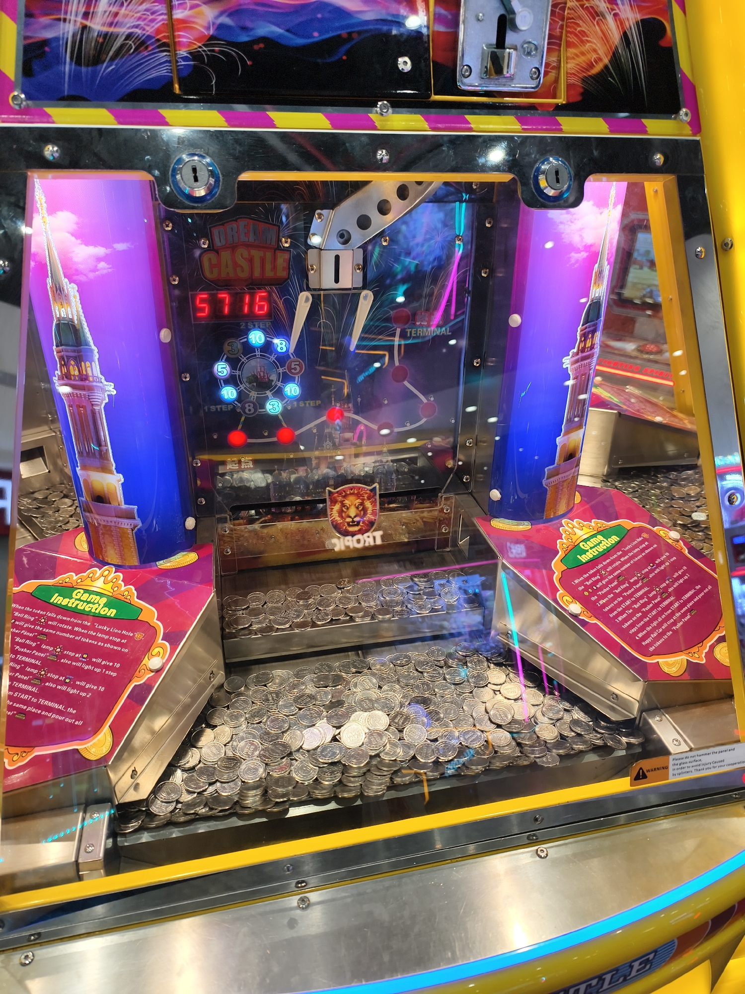 This image depicts a "Dream Castle" coin pusher arcade game. The machine is predominantly yellow with purple, blue, and pink accents. The game features a clear window showcasing a large quantity of silver-colored coins stacked on a moving platform. Above the coins are graphics of a castle tower and a display showing the number "5716." To the left and right of the central coin area are panels with game instructions printed on them. Warning labels are visible at the bottom of the machine. The location is an indoor arcade setting. The city and state are not defined.