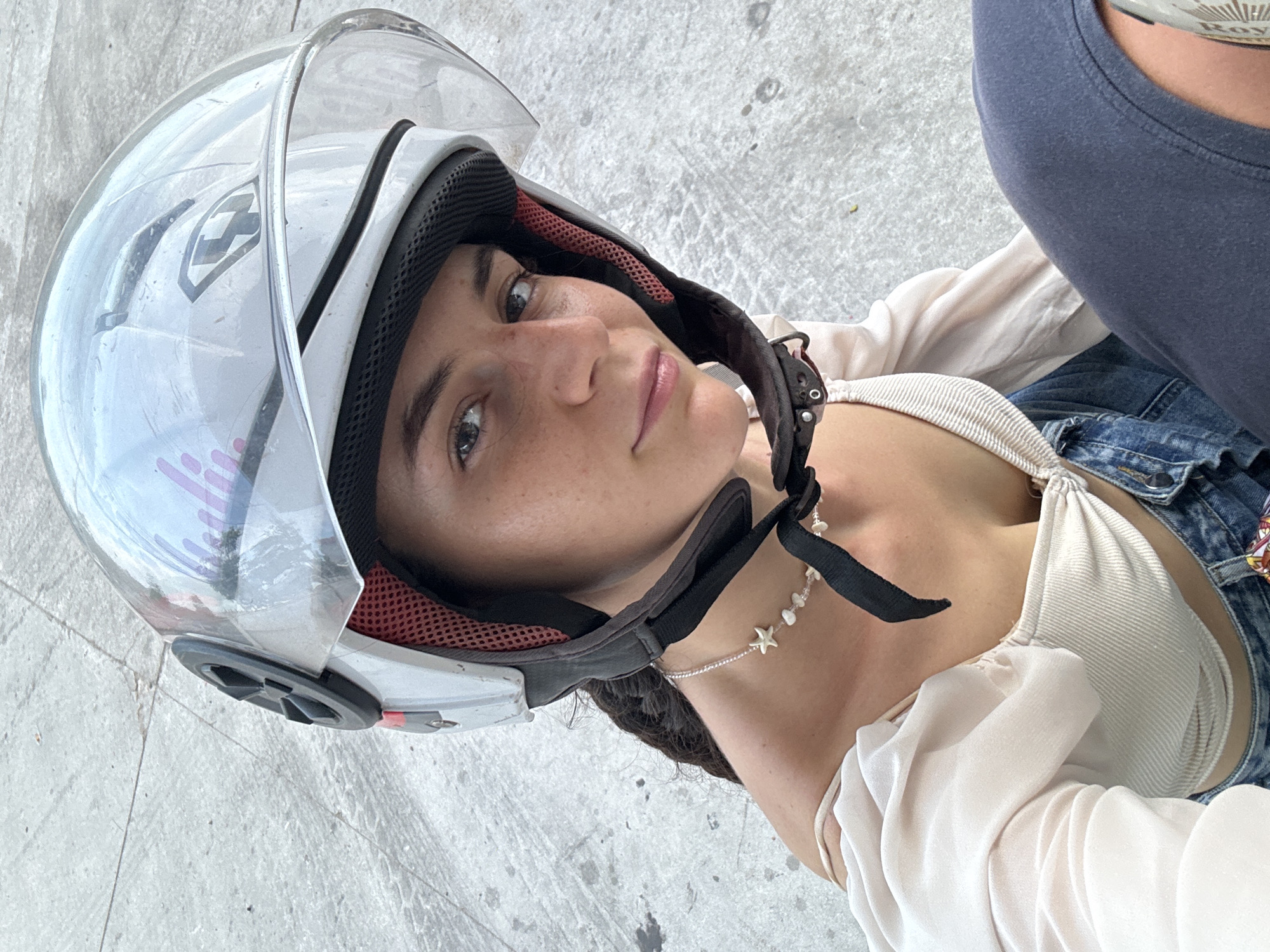 A woman wearing a white motorcycle helmet and a white top is looking at the camera. She is wearing a necklace with starfish pendants. Parts of a blue denim jacket and a dark gray shirt are also visible. The background is a gray concrete surface. The location is Phu Thuy Ward, Vietnam.
