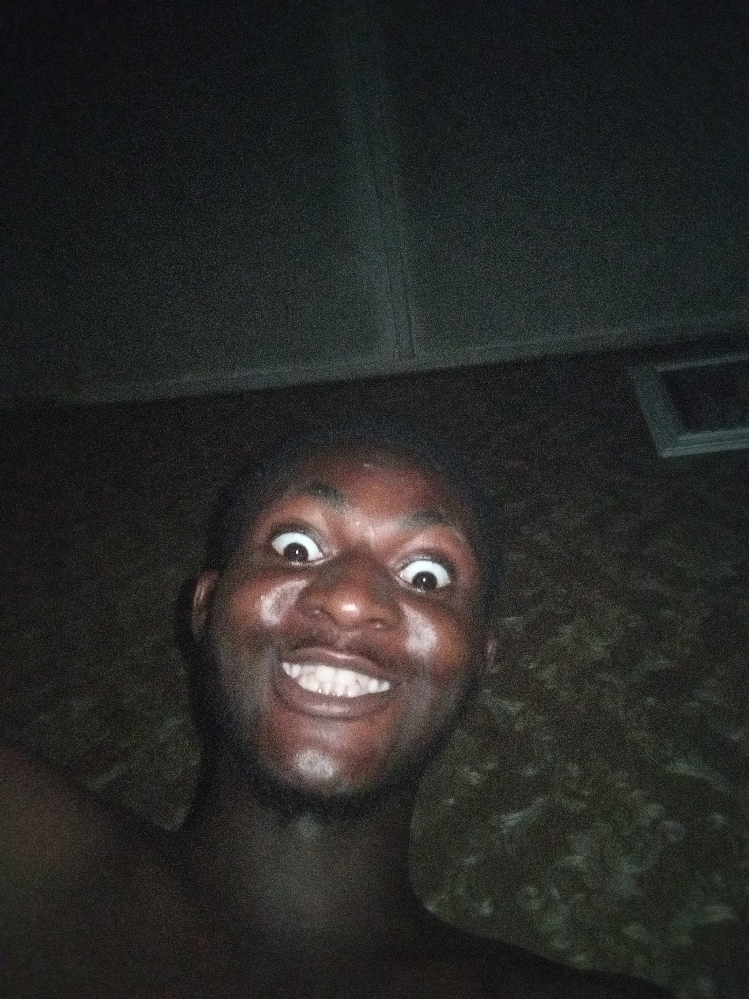 A close-up photograph depicts a dark-skinned man with wide eyes and a broad smile. He appears to be shirtless and is positioned at an upward angle to the camera. The background is dark, possibly a ceiling or wall with an indiscernible texture. A small rectangular object is partially visible in the upper right corner. The location is specified as Unknown, Unknown.
