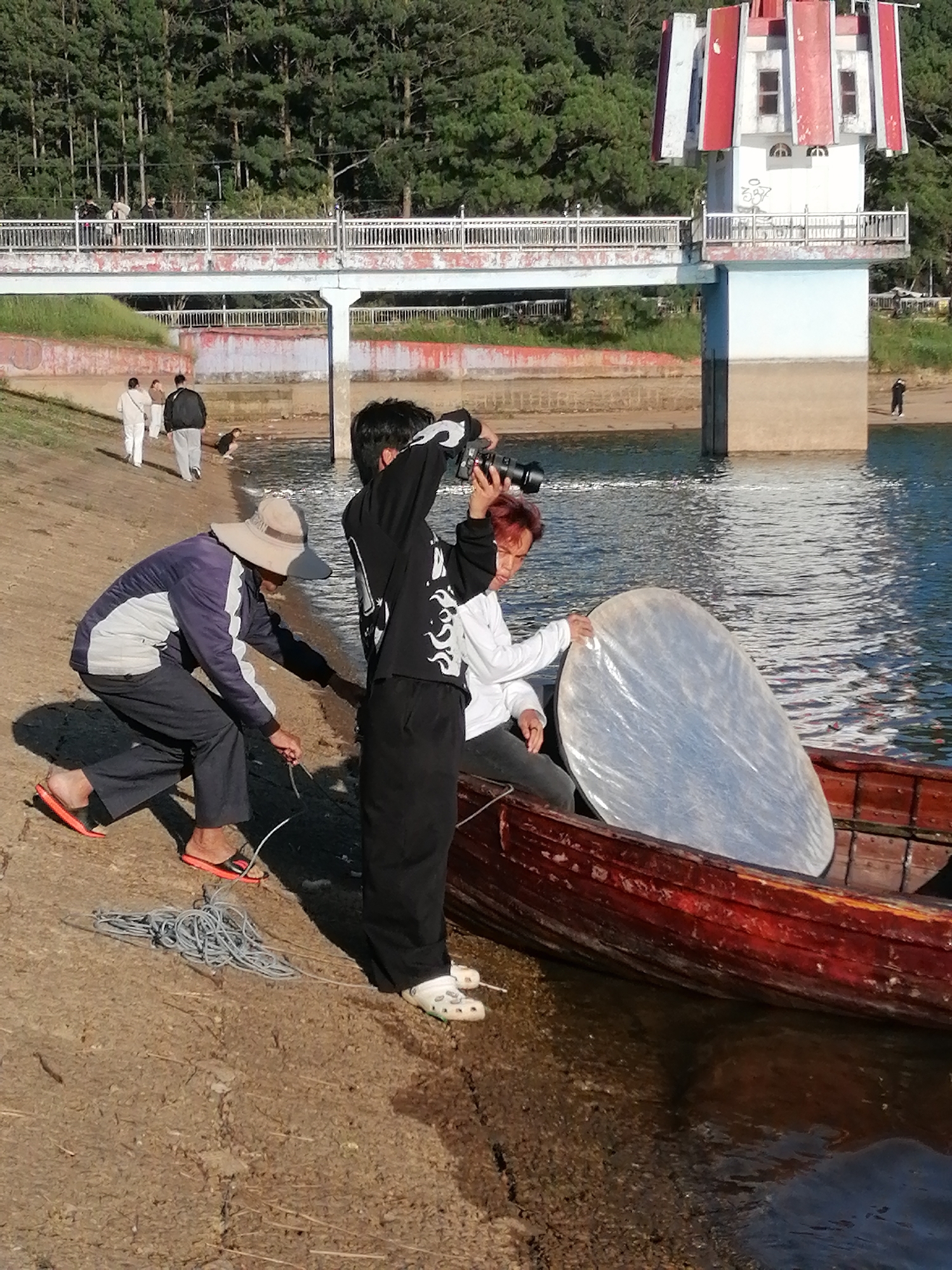The image depicts a daytime scene at Xuan Huong Lake in Da Lat, Vietnam. In the foreground, a man wearing a hat and dark pants is kneeling on the shore, appearing to be handling a coiled rope. Beside him, a person in a black long-sleeved shirt with a camera is standing and photographing something in a red wooden boat. Another person, wearing a white shirt and dark pants, is seated in the boat holding a large, round, reflective disc. The boat is partially submerged in the water. In the background, a bridge spans across the water, with several people walking on it. Further in the background, a distinctive tower with red and white accents stands next to the bridge. The surrounding landscape includes a dense forest of pine trees. The water of the lake is visible, reflecting the sky and the surrounding structures. The ground is a mix of sand and dirt.