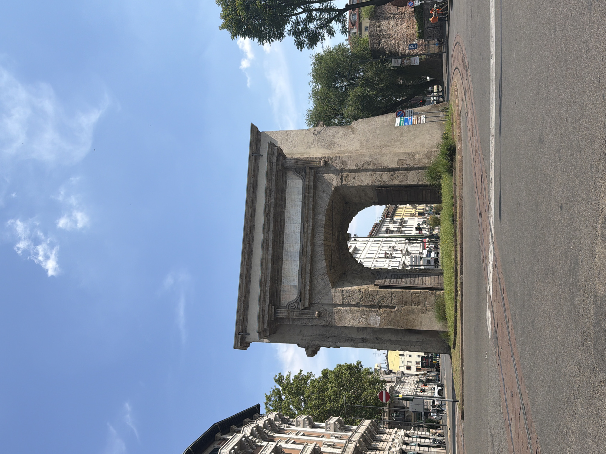 The image shows the Arco della Pace (Arch of Peace) in Milan, Italy, seen from a slightly low angle. The arch is a neoclassical monument made of stone. The sky is blue with some clouds. The street is to the right of the image. Behind the arch, there is a view of buildings. There are trees and road signs.