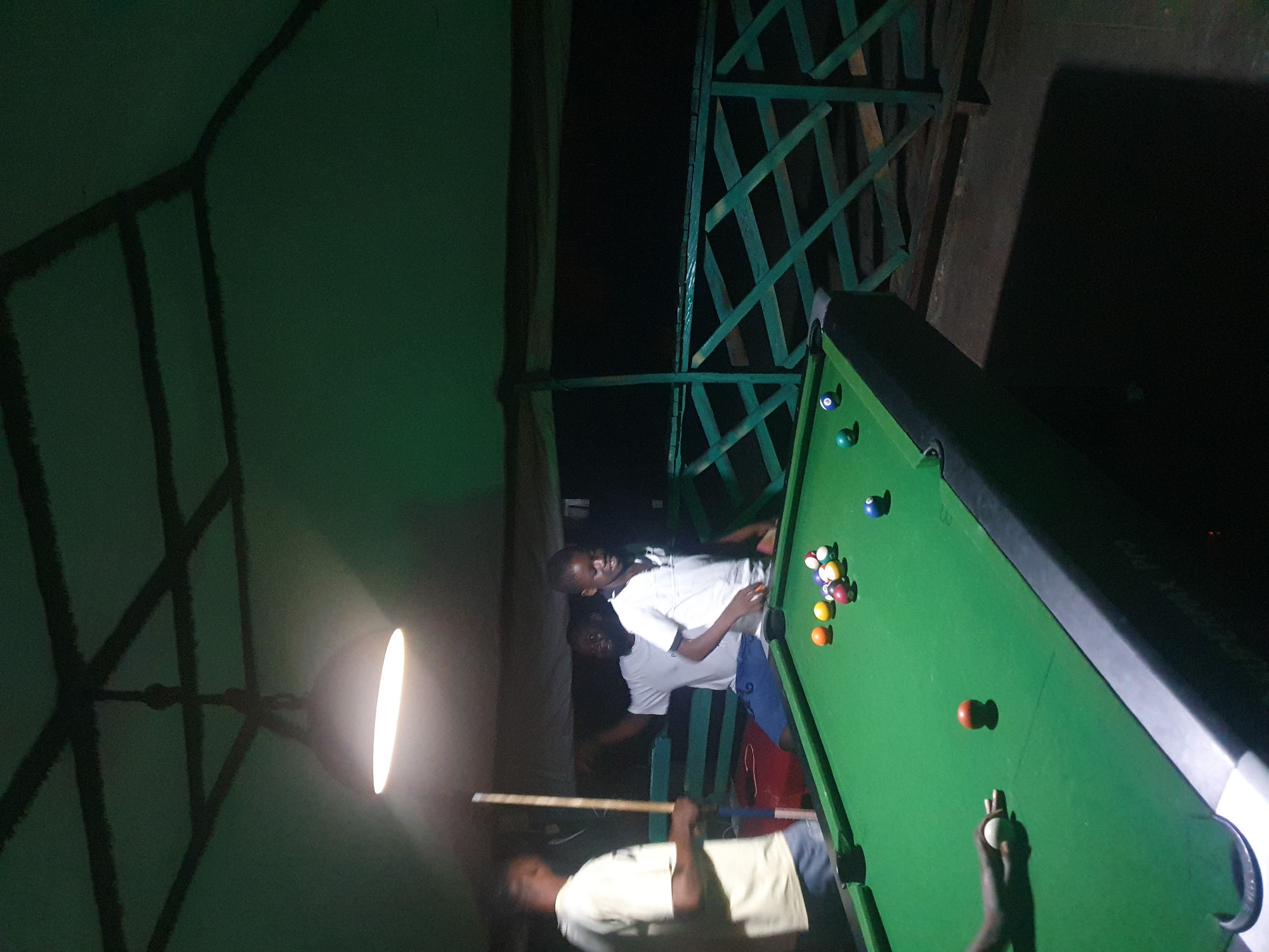 In Lokoja, Nigeria, an outdoor billiard hall has been set up under a partially covered structure with a green ceiling and a suspended lamp. Three individuals are visible around a green pool table. On the left, a person in a light-colored t-shirt and shorts stands holding a pool cue. In the center, a person in a white t-shirt and blue shorts appears to be interacting with another person. To the right, a person in a white t-shirt is looking at the pool table, which is covered with several billiard balls, including a white cue ball. The table's surface is green, with black cushions and pockets. A green railing with a criss-cross pattern forms the background of the scene. The lighting is dim, with the overhead lamp casting a bright glow. The ground around the table appears to be a dark, rough surface, possibly dirt or concrete.
