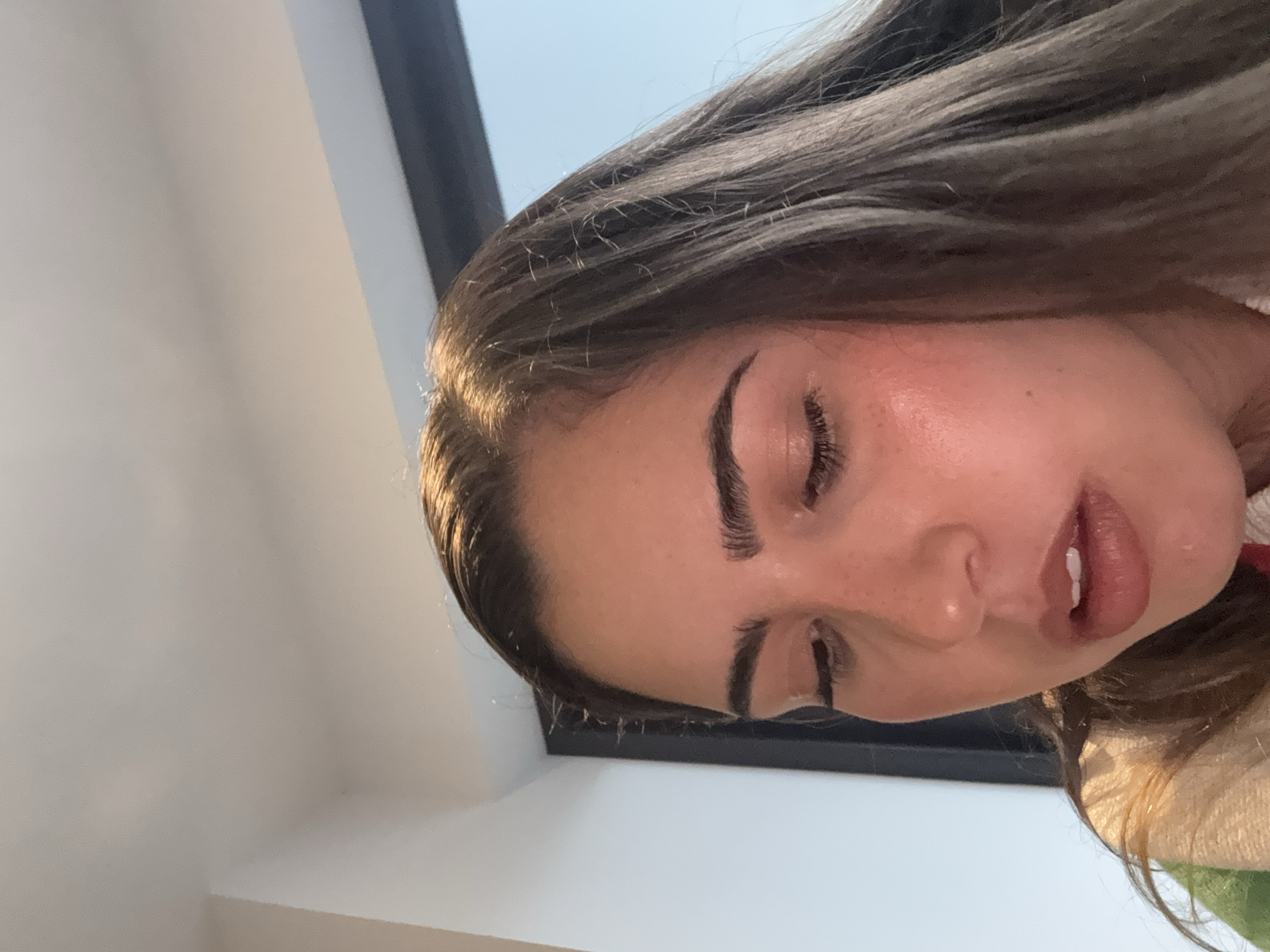 A close-up, angled view shows the face of a young woman with brown hair. Her eyes are closed. The background includes a white wall, a dark-framed window, and a glimpse of a green and white garment. The location within San Bernardino Tlaxcalancingo, Mexico is not discernible from the visual information.