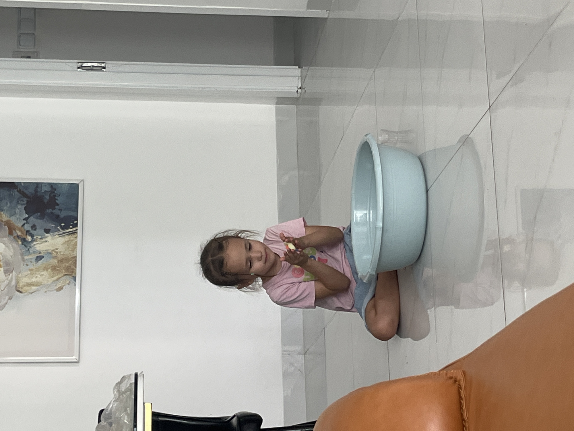 A young girl is kneeling on a white tile floor in what appears to be the city of Bac Nha Trang Ward, Vietnam. She is wearing a pink t-shirt and blue shorts and holding a yellow object in her hands, possibly playing with it. A large light blue plastic bowl is placed near her. The background includes a white wall with a painting and what seems to be a doorway with a metal lock. A brown leather couch is partially visible in the foreground.