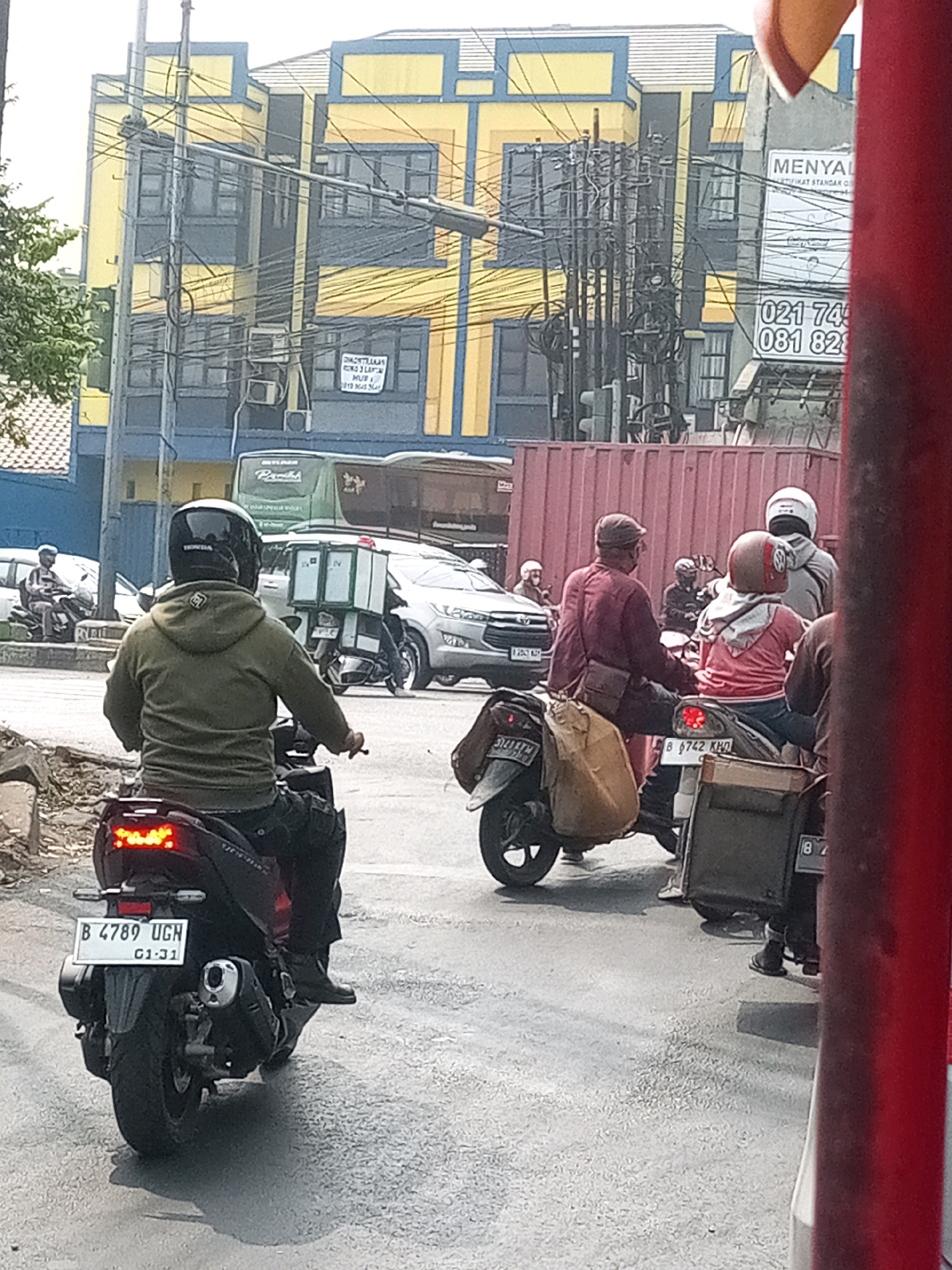 The image depicts a street scene in Bekasi, Indonesia, likely during the daytime. In the foreground, a person is riding a dark-colored scooter, their back to the camera. They are wearing a green hooded jacket and dark pants, with a black helmet. The scooter has a license plate reading "B 4789 UGH" with "GI-31" below it. The rear brake lights of the scooter are illuminated. To the right of the foreground scooter, another scooter is visible, carrying two people, one wearing a pink top and the other a gray top and helmet. This scooter has a license plate "B 6742 KHD". In the background, several cars and more scooters are present, along with a large red shipping container. Buildings with yellow and blue facades line the street, adorned with numerous electrical wires. A green bus with "Pramitha" written on it is also visible. The road appears wet in places, suggesting recent rain.