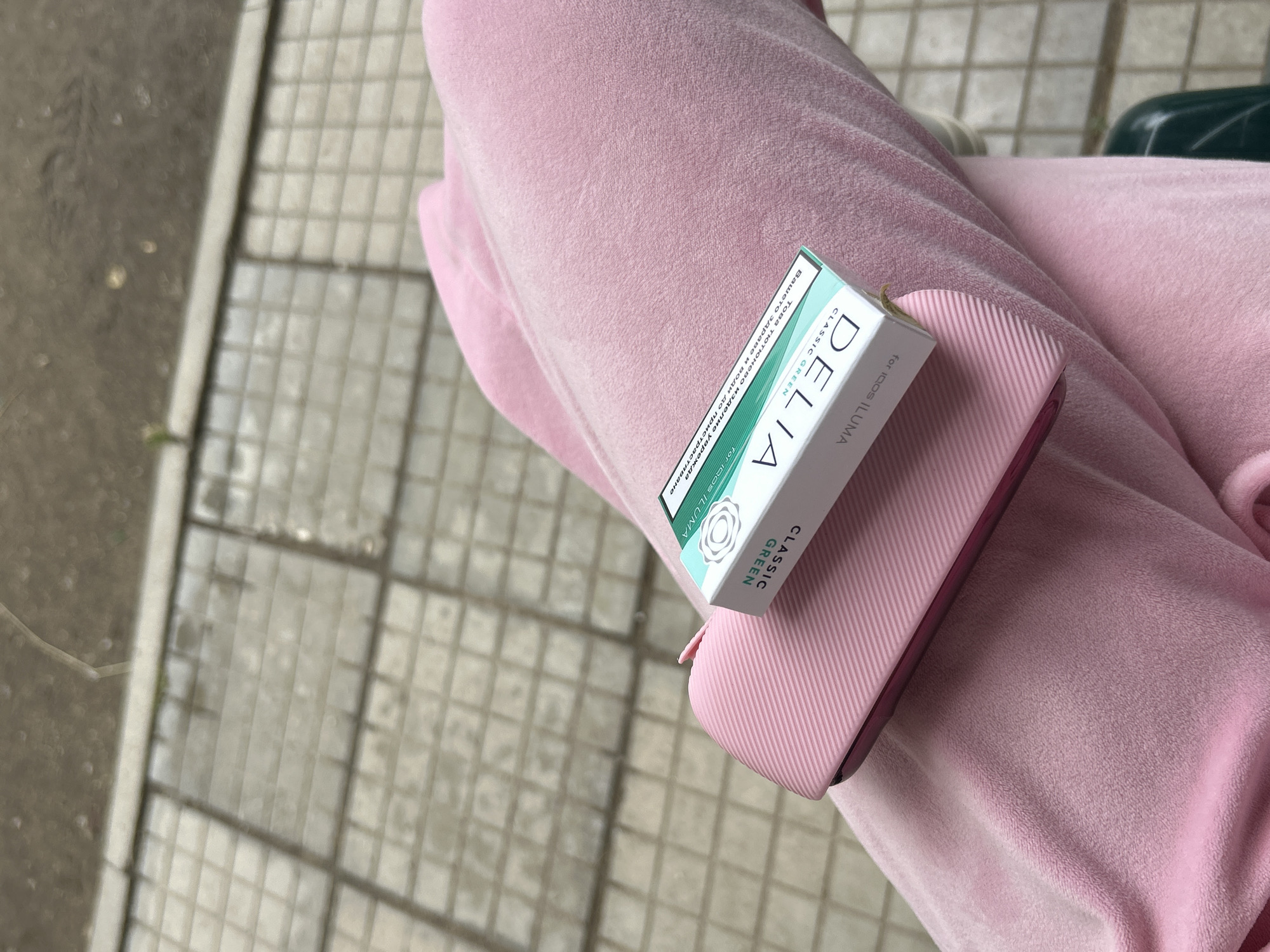 A close-up, high-angle shot captures a light pink, possibly velvet or plush fabric, which appears to be clothing, likely on a person's lap or leg. Resting on this fabric are two objects: a textured, light pink case or device holder, and a small, rectangular box with white and green coloring.
The box prominently displays the brand name "DELIA" and "CLASSIC GREEN," with the additional text "for IQOS ILUMA." On one side of the box, a health warning is visible in Cyrillic script: "Вашето здраве и води до пристрастяване Това тютюнево изделие уврежда" (Your health and leads to addiction. This tobacco product harms). The light pink case has a ribbed or grooved texture on its visible surface.
The background shows a paved surface, likely a sidewalk or outdoor path, with a pattern of light grey square tiles on the right and a darker, rougher ground surface on the left, separated by a light-colored line, possibly a curb or edge. The lighting suggests daytime, possibly overcast, with no harsh shadows. The scene implies a casual outdoor setting. The presence of Bulgarian health warnings aligns with the context of Plovdiv, Bulgaria.