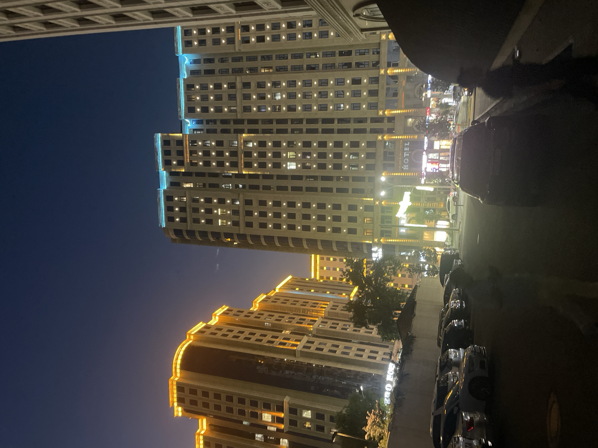 The image depicts a nighttime scene in Dushanbe, Tajikistan. The primary subjects are two modern high-rise buildings, illuminated by warm yellow and cool blue lights, suggesting an urban environment. The buildings are densely packed with windows, many of which are lit from within, indicating occupancy.
In the foreground, a row of parked cars is visible along a street. The cars appear to be modern sedans and SUVs, suggesting a level of affluence. The streetlights cast a dim glow on the vehicles and the pavement. The overall lighting suggests it is nighttime, likely after sunset.
A prominent sign on one of the buildings reads "HOTEL" in capital letters, identifying the function of at least part of the structure. Other signs with text are also visible but are too blurry to be deciphered. The scene is calm and quiet, with no visible people or active events taking place. The weather appears clear, as the sky is dark and the lights are sharp. The architecture of the buildings, with their clean lines and extensive glass facades, suggests a contemporary urban setting. The image conveys a sense of nighttime urban life, with the buildings and cars representing the city's infrastructure and modern development.