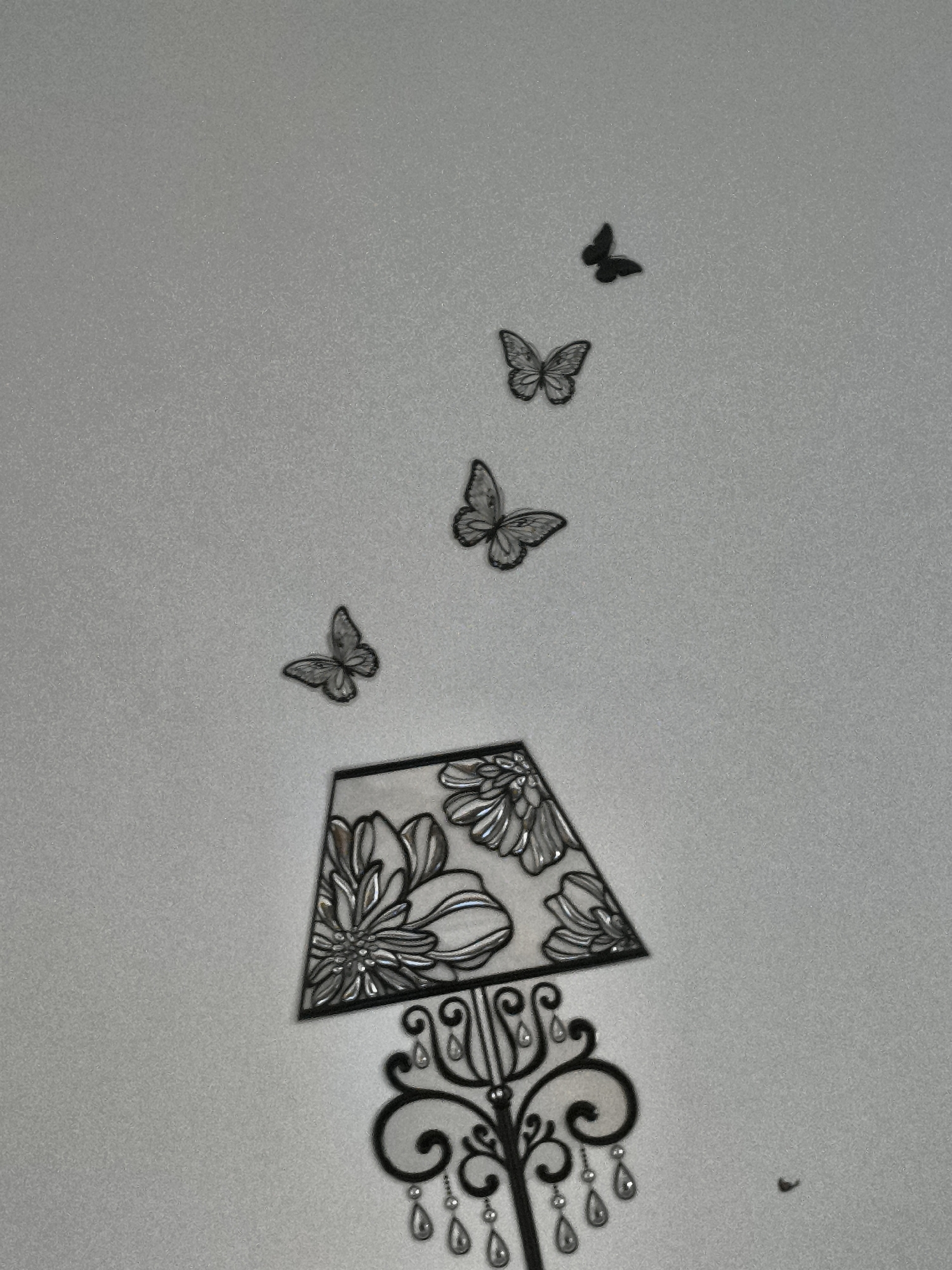 A wall with a decorative lamp and butterflies. The lamp is a dark color with a floral pattern on the shade and a curvy, ornate stand with teardrop-shaped accents. Four butterflies are positioned above the lamp, seemingly "flying" upwards. One additional butterfly is located off to the side of the lamps base. All elements are silhouettes or appear to be flat wall decals. The wall itself is a pale, possibly off-white color. It is unlikely the image is located in Pélussin, France, as it is simply a photo of a wall decoration.