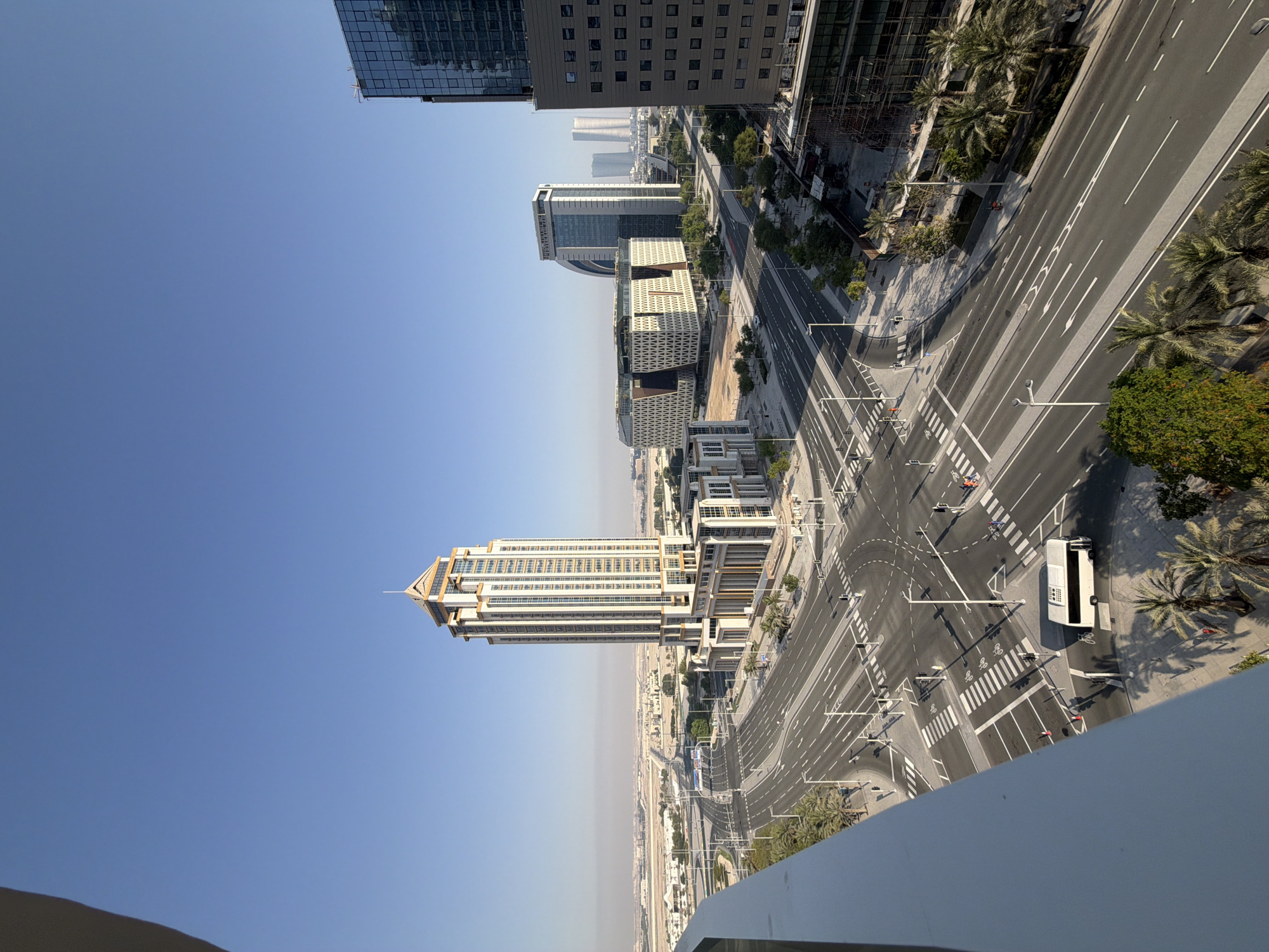 This is a high-angle, full shot of a wide, multi-lane road intersection in a modern city. The sky is clear and blue with a slight haze in the distance. On the right side of the image, a tall, modern office building with a glass facade is visible. To the left of this building, further down the road, is a distinctively patterned, angular building with white and brown geometric designs. A very tall, cream-colored skyscraper with a peaked roof stands prominently in the middle ground. In the foreground, a white railing from a balcony or building overhang partially obscures the left side of the image.
The road is paved with asphalt and marked with white lines, including crosswalks and directional arrows. A white bus is stopped at the intersection, and a few pedestrians are visible crossing the street. Palm trees are scattered along the roadside, and some greenery is present in landscaped areas. In the background, a few other modern buildings can be seen, along with what appears to be undeveloped land or construction sites. The overall impression is one of a bustling urban environment with contemporary architecture and infrastructure.
