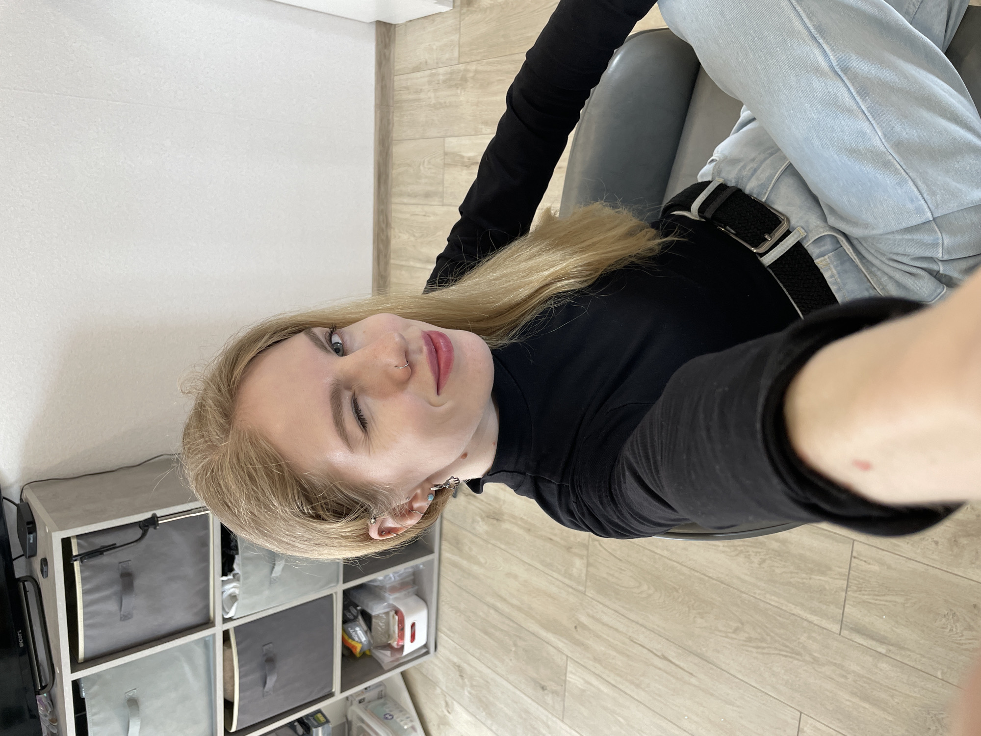 An interior shot depicts a young adult female seated in a grey chair, taking a selfie. She has long blonde hair, light-colored eyes, and is winking with her right eye while looking directly at the camera with a subtle smile. She wears a black long-sleeved turtleneck shirt, light blue jeans, and a black belt with a silver buckle. Her left nostril features a silver ring piercing, and her left ear displays multiple silver piercings, including one with a small charm resembling a Hamsa hand. A small red mark is present on the inner forearm of her extended left arm, which holds the camera. The background reveals a light-colored cube storage unit with grey fabric bins to the left. A dark flat-screen television rests on top of the unit, accompanied by a small black electronic device. The room features light-colored laminate flooring and plain white walls. No specific identifying features of Izhevsk, Russia, are discernible within the image.