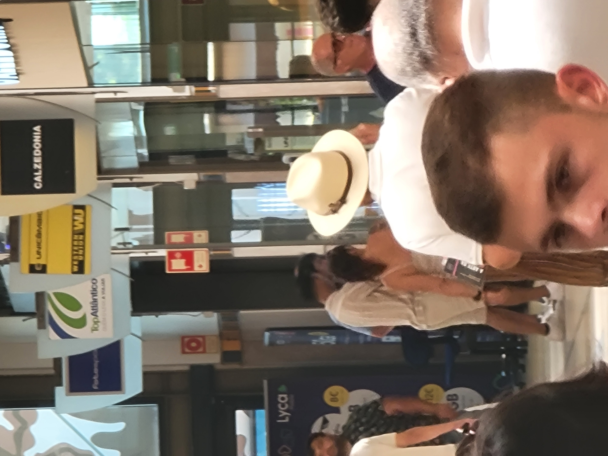 The image depicts an indoor scene, possibly a shopping mall or an airport, with several individuals visible. In the foreground, the back of a man wearing a white shirt and a light-colored fedora is prominent. To his right, the side of another man with gray hair and sunglasses is visible. Behind them, a couple is seen in mid-conversation; the woman is wearing a light-colored dress, and the man is in a white shirt and shorts. Further back, a man with a beard wearing a patterned shirt is seen. Above, there are several signs, including one for "CALZEDONIA," "TopAtlântico," and "Lyca Mobile." The overall environment appears to be a public indoor space with various shops or service points. The presence of these commercial signs suggests a commercial area. The setting is likely a public indoor space within the city of Faro, Portugal, given the "TopAtlântico" sign which often indicates services related to tourism or travel in Portugal.
