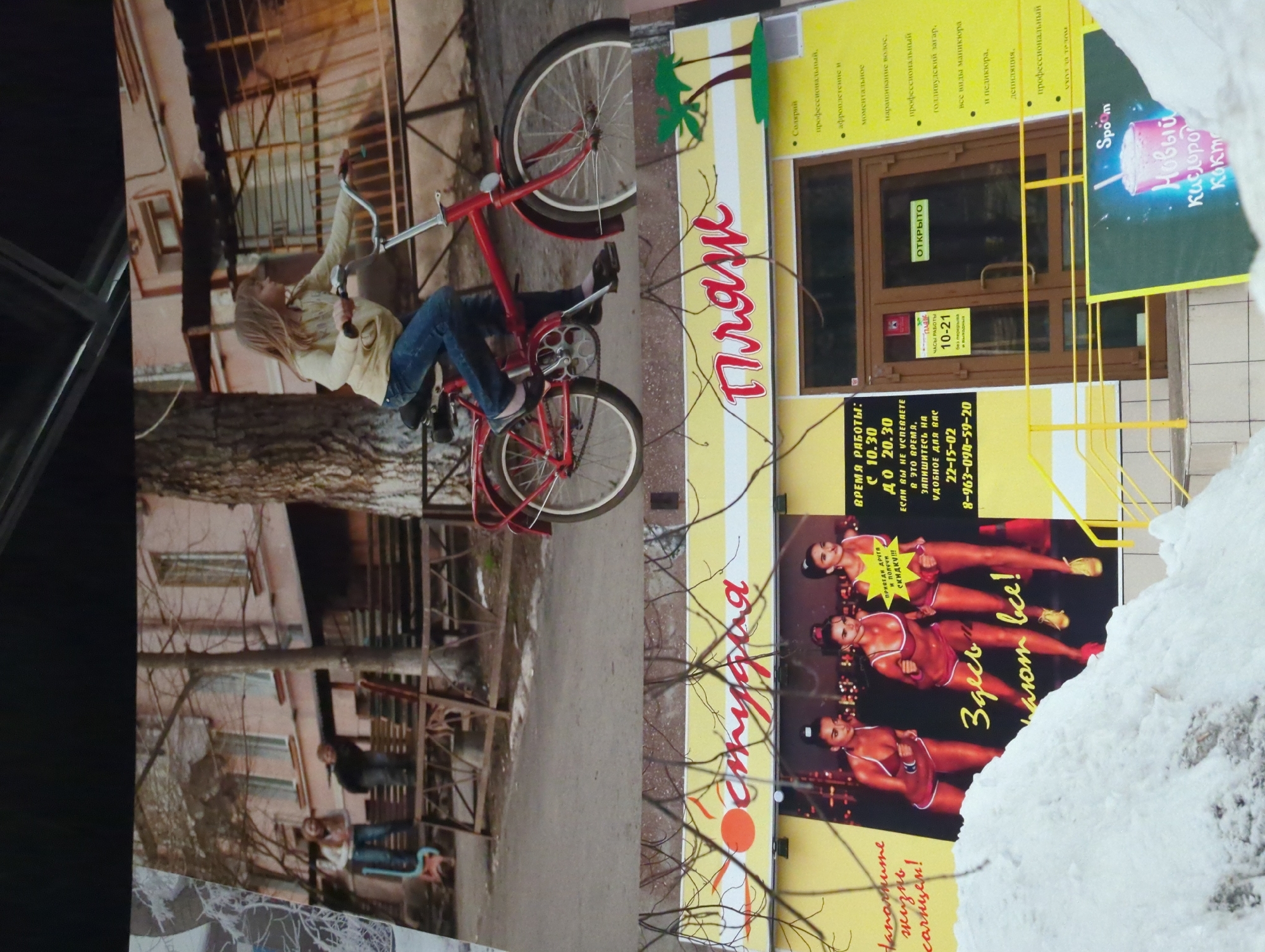 The image displays a street scene featuring a red bicycle with a young girl riding it, and in the background, a building with a sign that reads "Студия Пляж" which translates to "Studio Beach". The sign also features images of three women in athletic wear, and text that appears to be advertisements for services. One part of the sign indicates "HOURS OF OPERATION: 10-21". Another section of the sign advertises a "New Oxygen Cocktail". The foreground shows a pile of snow, suggesting a cold weather environment. The building appears to be a commercial establishment. There is no indication of the location being in Jinniu District, China.