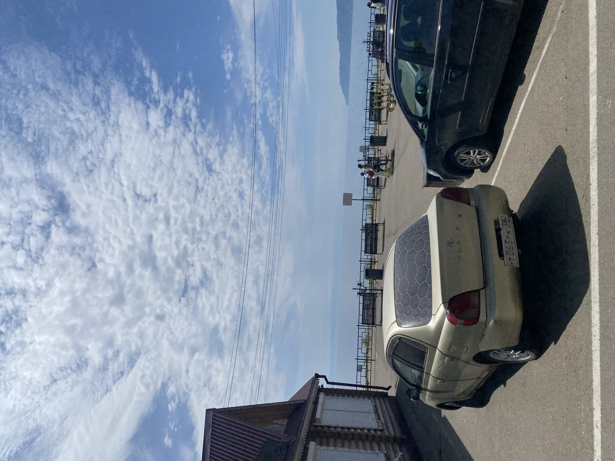 In Listvyanka, Russia, a sunny day unfolds with a view of a vast body of water, likely Lake Baikal, under a partially cloudy sky. A wooden building with a red corrugated roof and white shutters stands on the left, adjacent to a paved parking area. Two cars are parked: a light-colored sedan in the foreground, its rear window decorated with a hexagonal pattern, and a dark-colored SUV to its right. Beyond the parking lot, a metal fence lines a promenade with several benches and trash receptacles. In the distance, a few people are visible walking, and some yellow electric scooters are parked. The water stretches to the horizon, where a hilly coastline is faintly discernible. Power lines crisscross the sky above the scene.