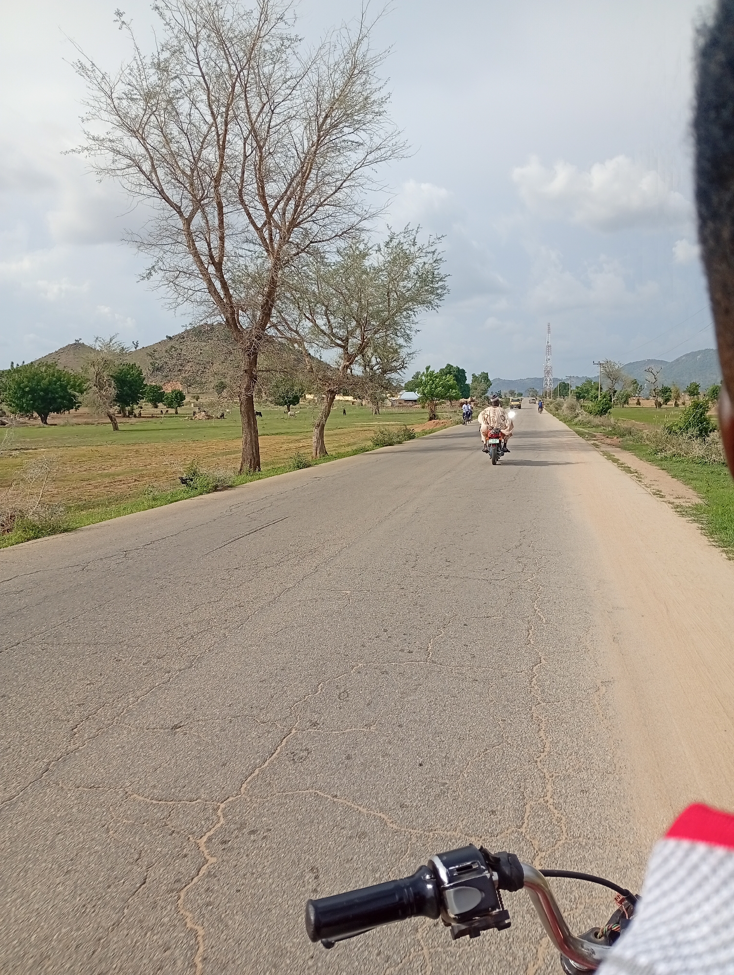 The image depicts a scene on a rural road in Shuwa, Nigeria. The perspective is from a vehicle, likely a motorcycle, with the handlebars visible in the foreground. The road is paved and shows signs of wear and tear, with cracks visible on the surface. The environment is characterized by dry, sparse vegetation and a few trees with bare branches. In the distance, a hill or mountain range can be seen, and there are some buildings or structures scattered across the landscape. Several people are present on the road, some riding motorcycles and others possibly walking. The sky is partly cloudy. The overall impression is of a dry, somewhat desolate rural area.